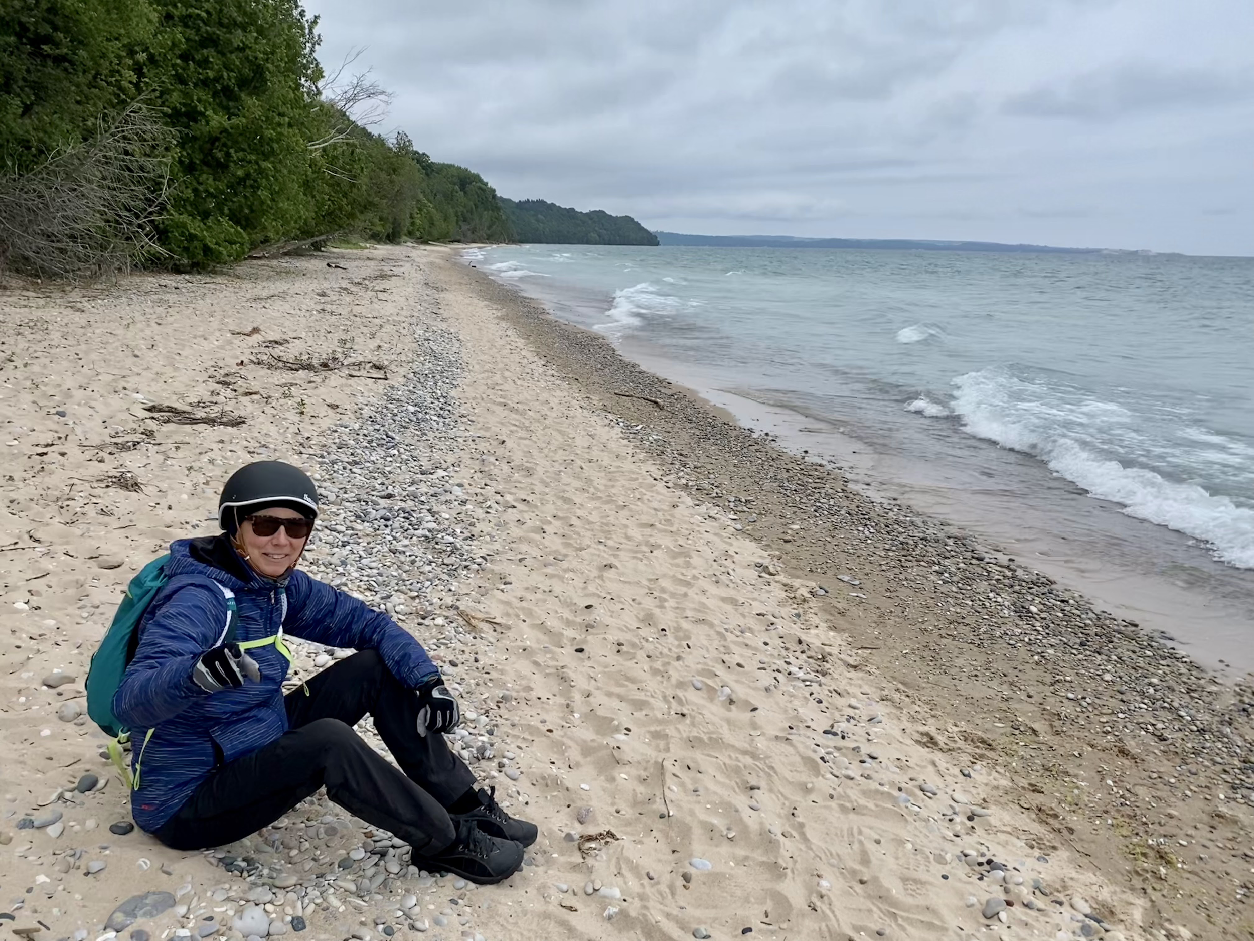Person sitting on a sandy beach wearing a helmet, sunglasses, and a blue jacket with a backpack, overlooking the ocean with trees and cloudy sky in the background.