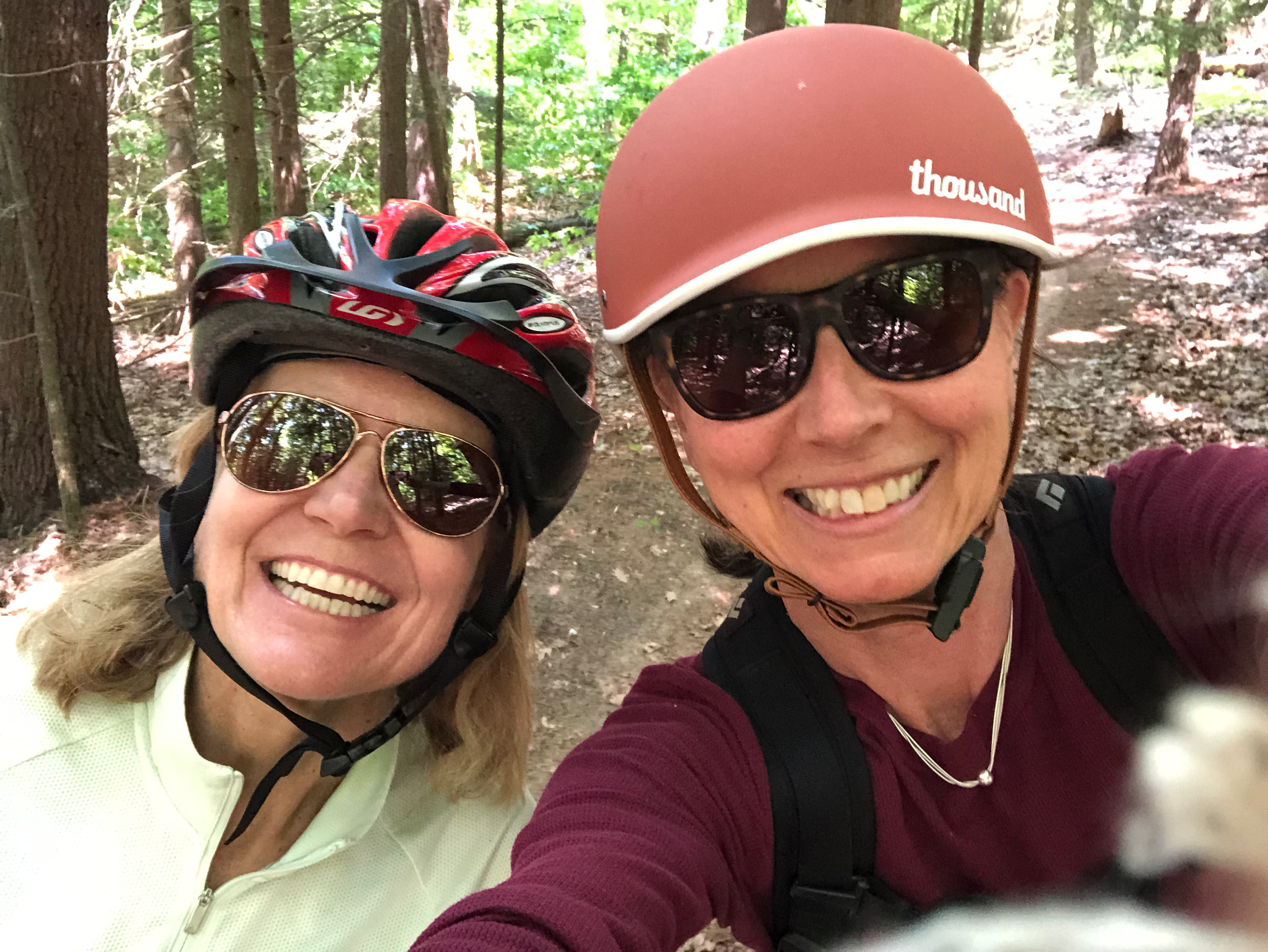 Two women wearing helmets and sunglasses taking a selfie in a wooded area during a bike ride.