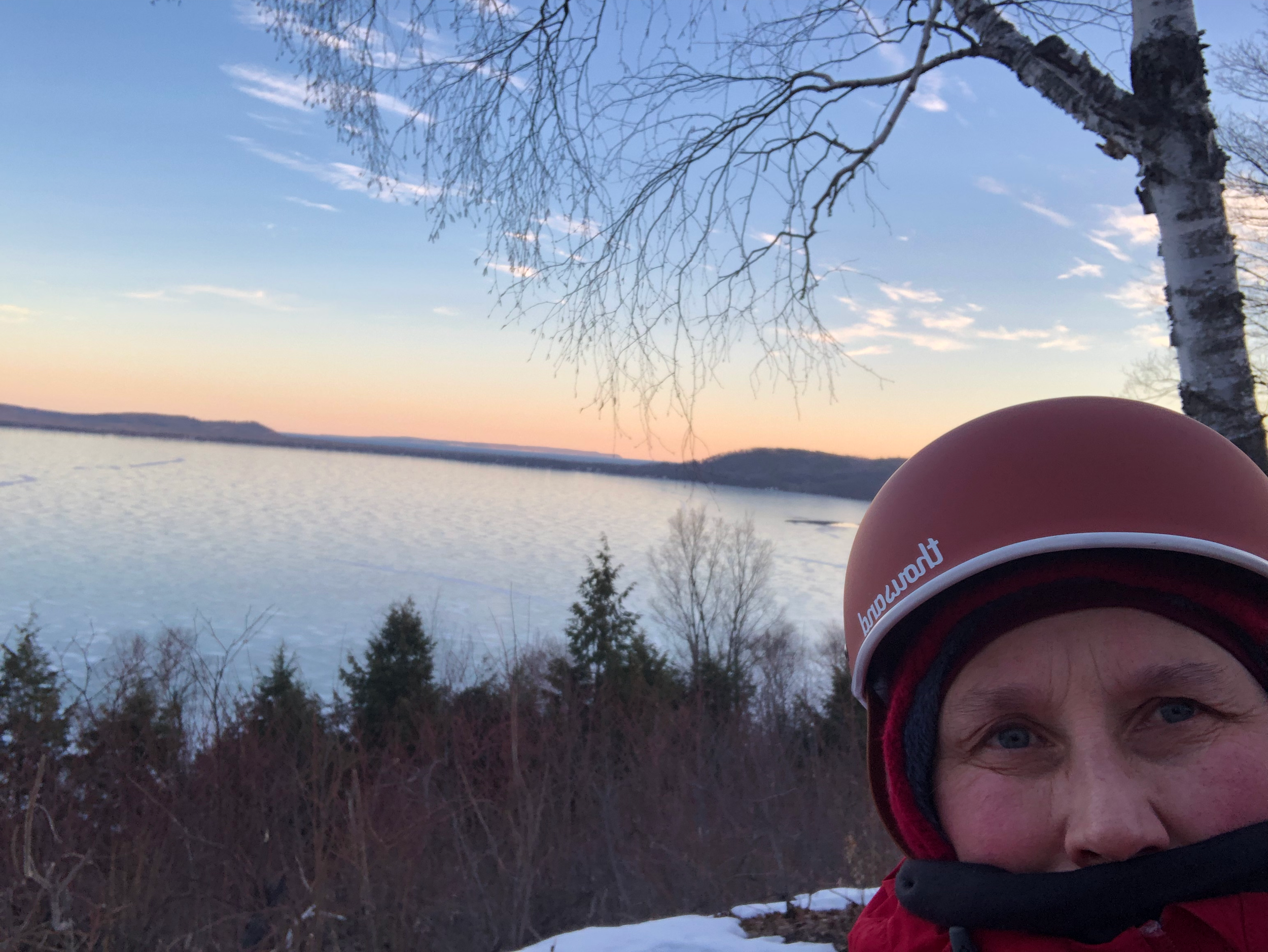 A woman wearing a red helmet and winter clothing taking a selfie outdoors near a snow-covered landscape with a lake, trees, and a sunset sky in the background.