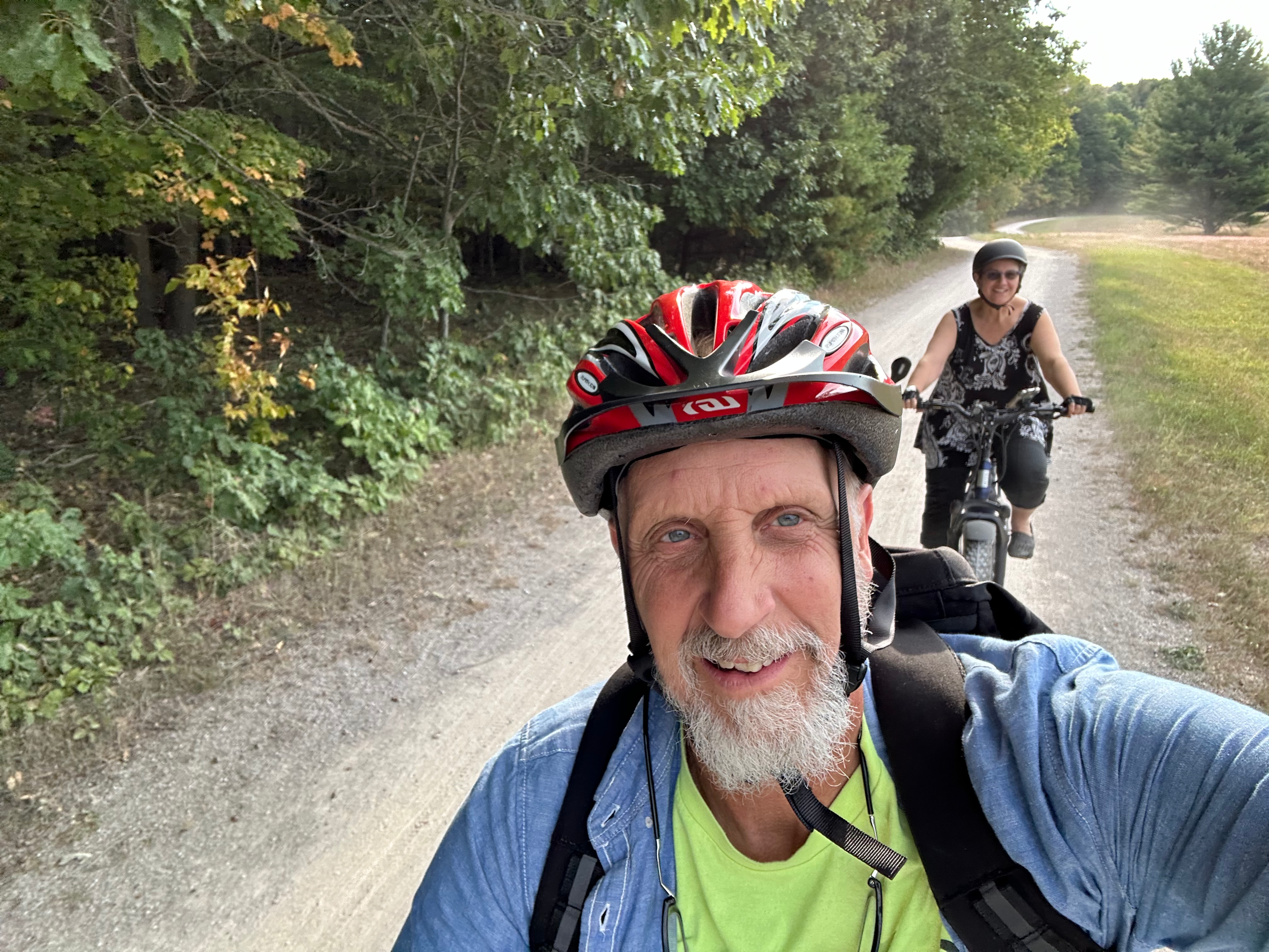 Two people riding bicycles on a gravel trail through a wooded area. The man in the foreground is wearing a red bicycle helmet, a green shirt, and a blue jacket, smiling at the camera. The woman in the background is wearing a helmet and sunglasses, pe