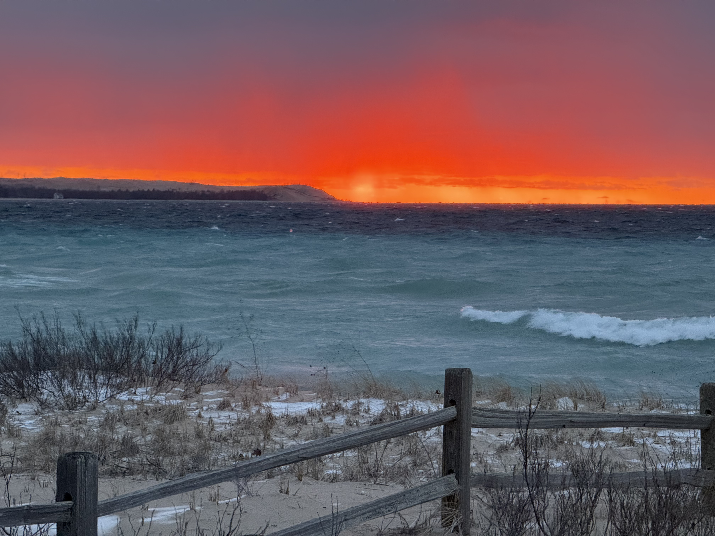 A sunset over the ocean with orange and purple sky, turbulent waves, and a sandy beach with sparse vegetation and a wooden fence in the foreground.