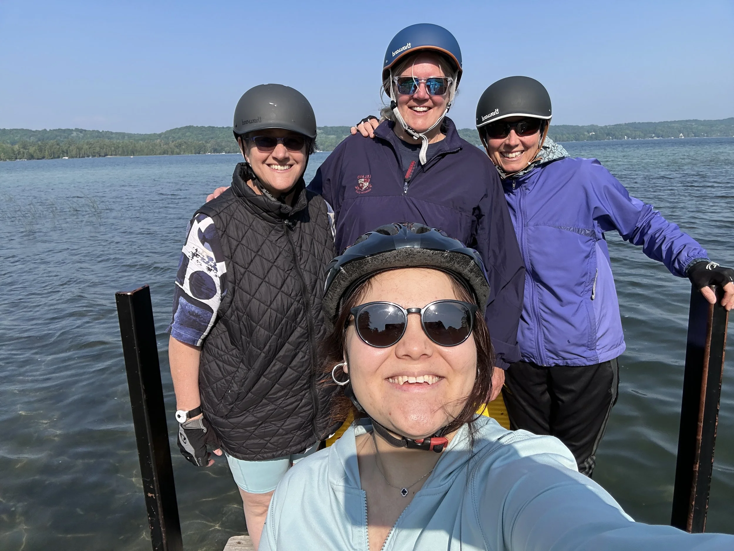 Five women wearing helmets, sunglasses, and outdoor clothing taking a group selfie on a boat with a lake and forest in the background.