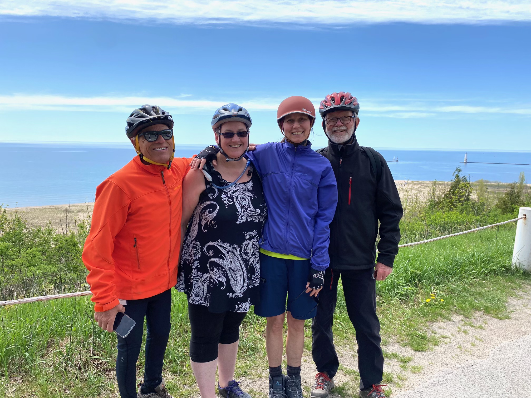 Four people standing outdoors, wearing helmets and athletic clothing, with a scenic ocean view and lighthouse in the background.