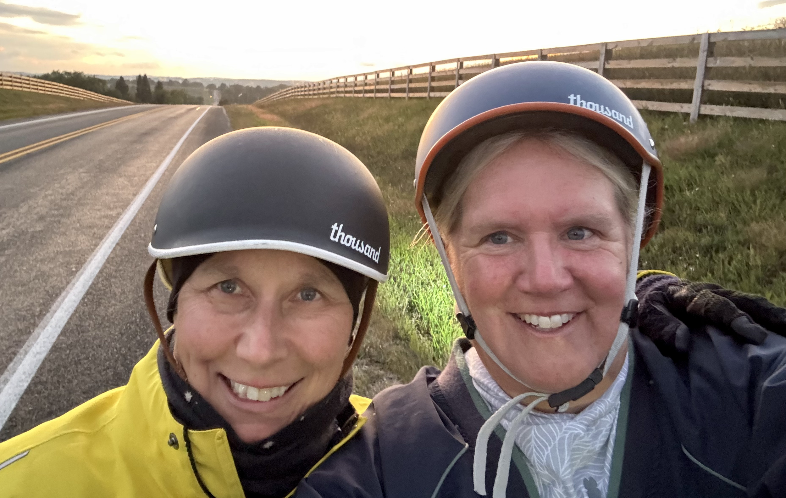 Two smiling people wearing helmets and jackets taking a selfie on a rural road at sunset, with grassy fields and a wooden fence in the background.