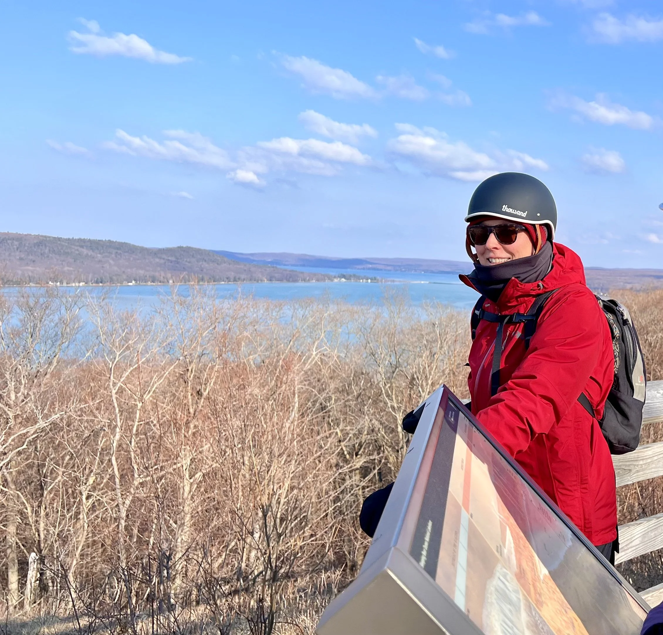 A person wearing a black helmet, red jacket, sunglasses, and a backpack smiling while sitting on a wooden railing overlooking a lake with hills in the background and a partly cloudy sky.