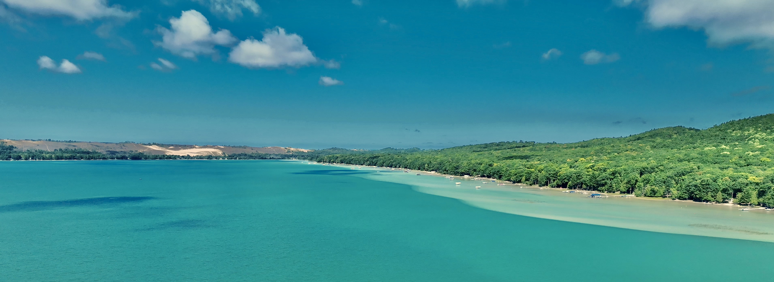Panoramic view of a turquoise lake with a sandy beach, lush green forest along the shoreline, and hills in the background under a partly cloudy sky.