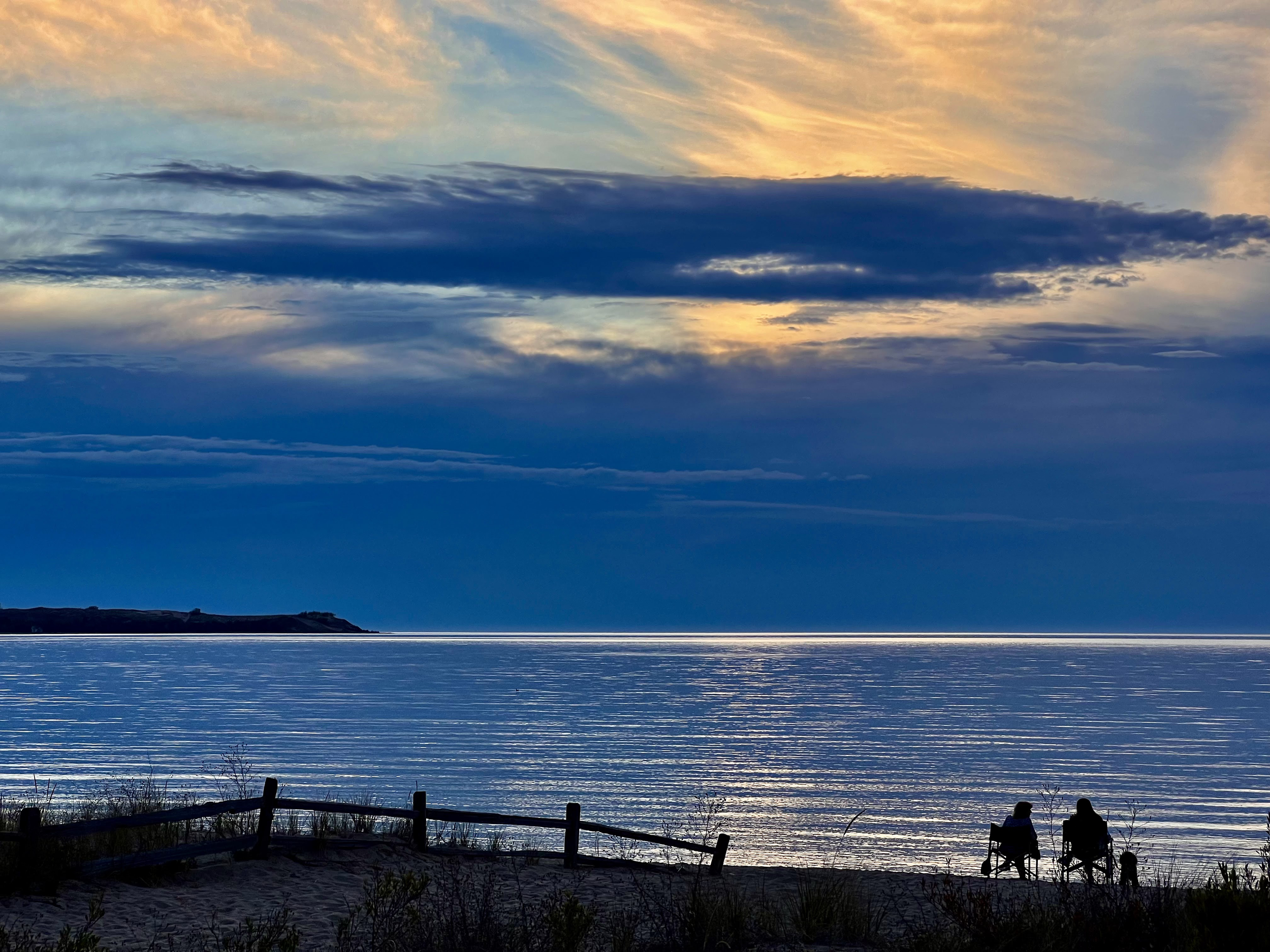 Two people sitting on chairs at the beach during sunset, overlooking calm water with a cloudy sky and a distant landmass.