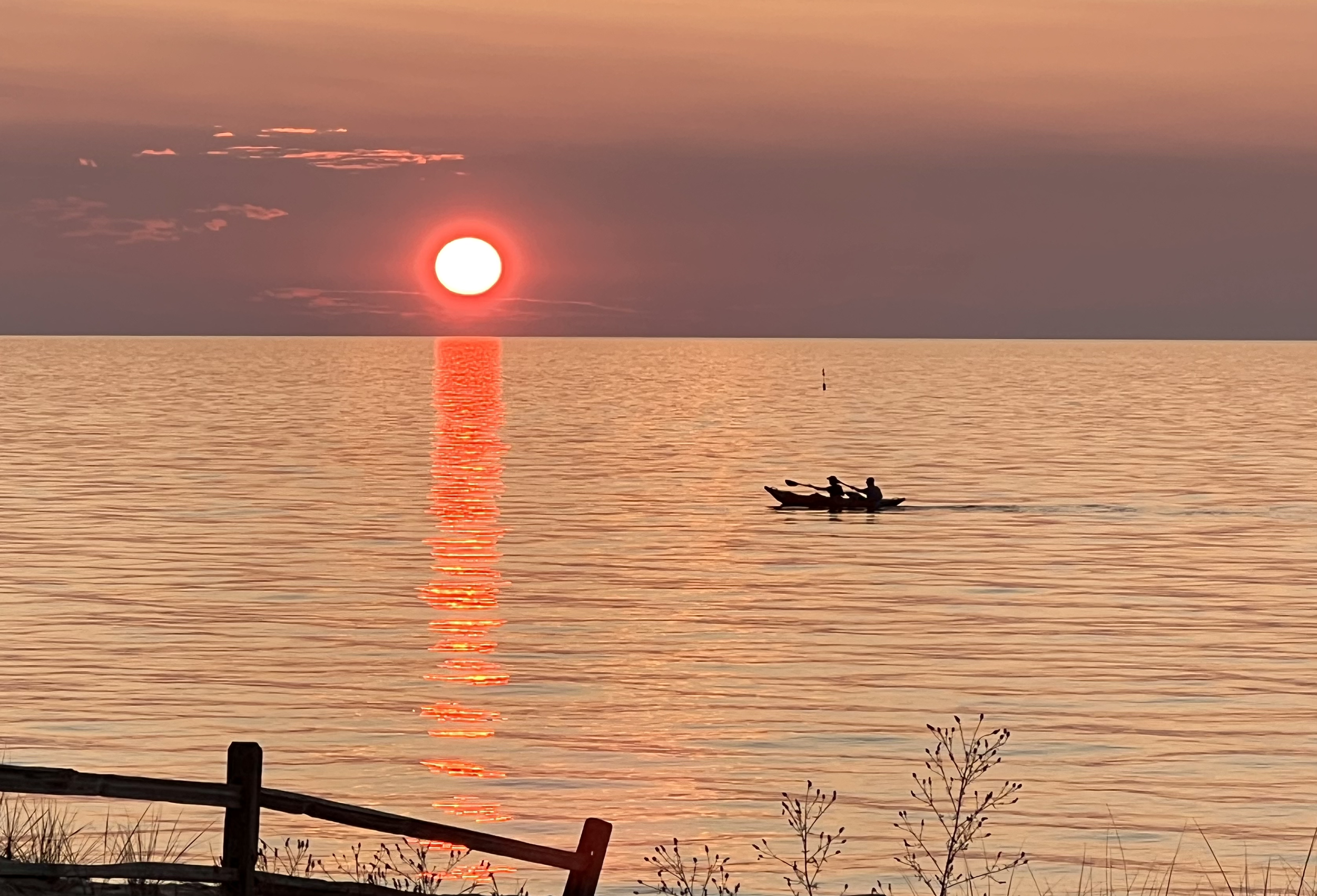 Sunset over the ocean with a small boat carrying two people and a wooden fence in the foreground.