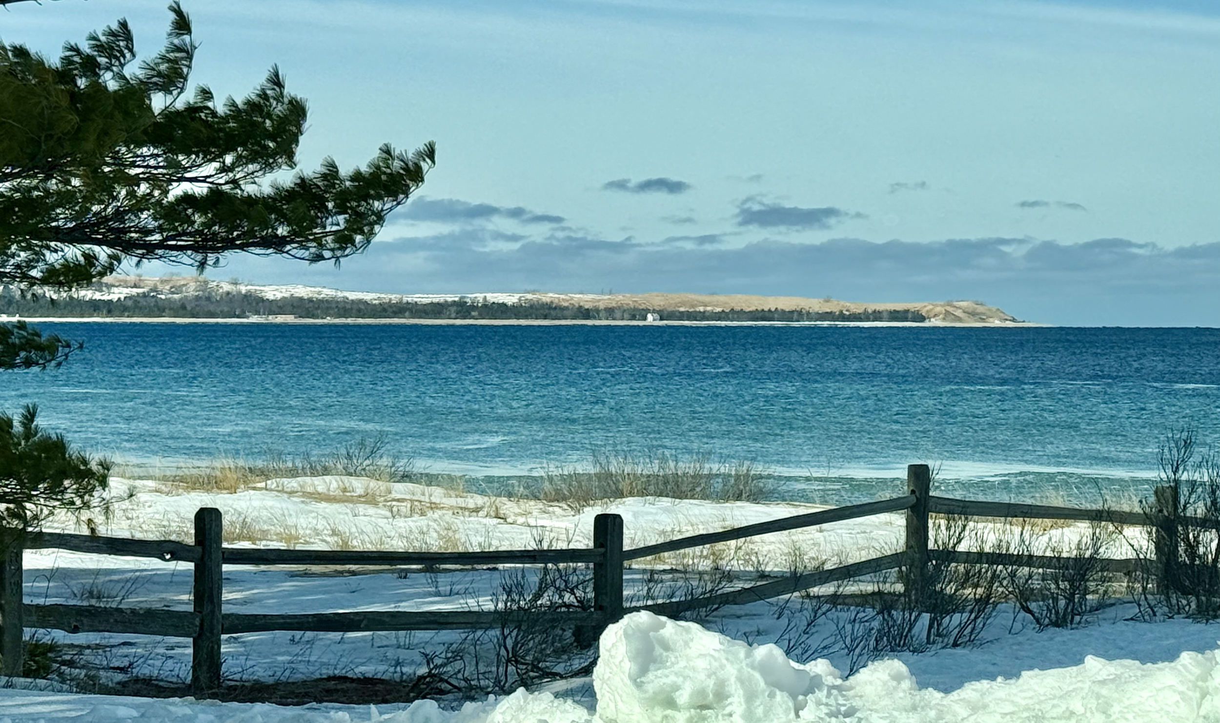 Snow-covered beach with a wooden fence, evergreen trees, blue water, and a distant land across the water under a partly cloudy sky.