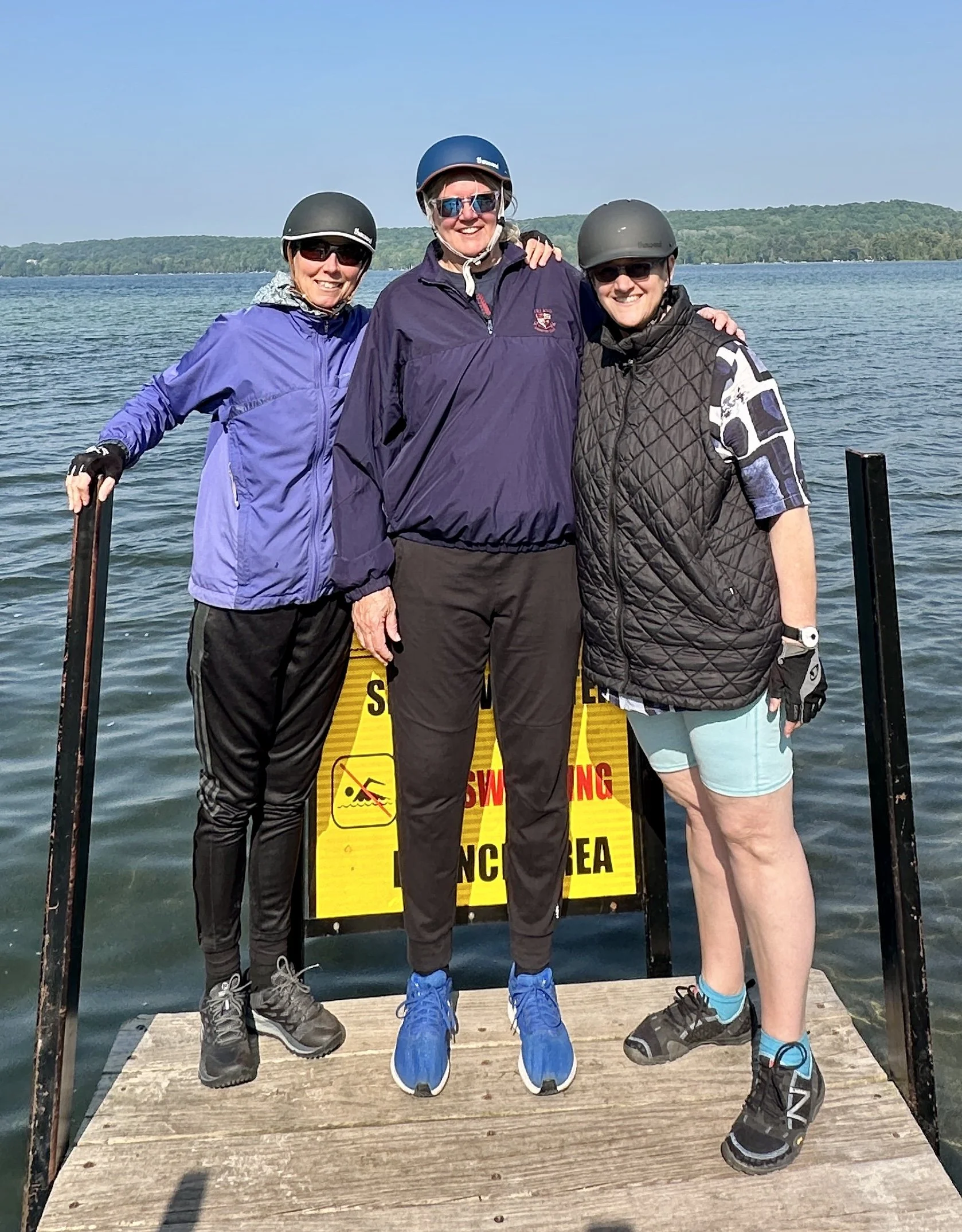 Three women standing on a dock by a lake, wearing helmets and outdoor clothing, smiling at the camera.