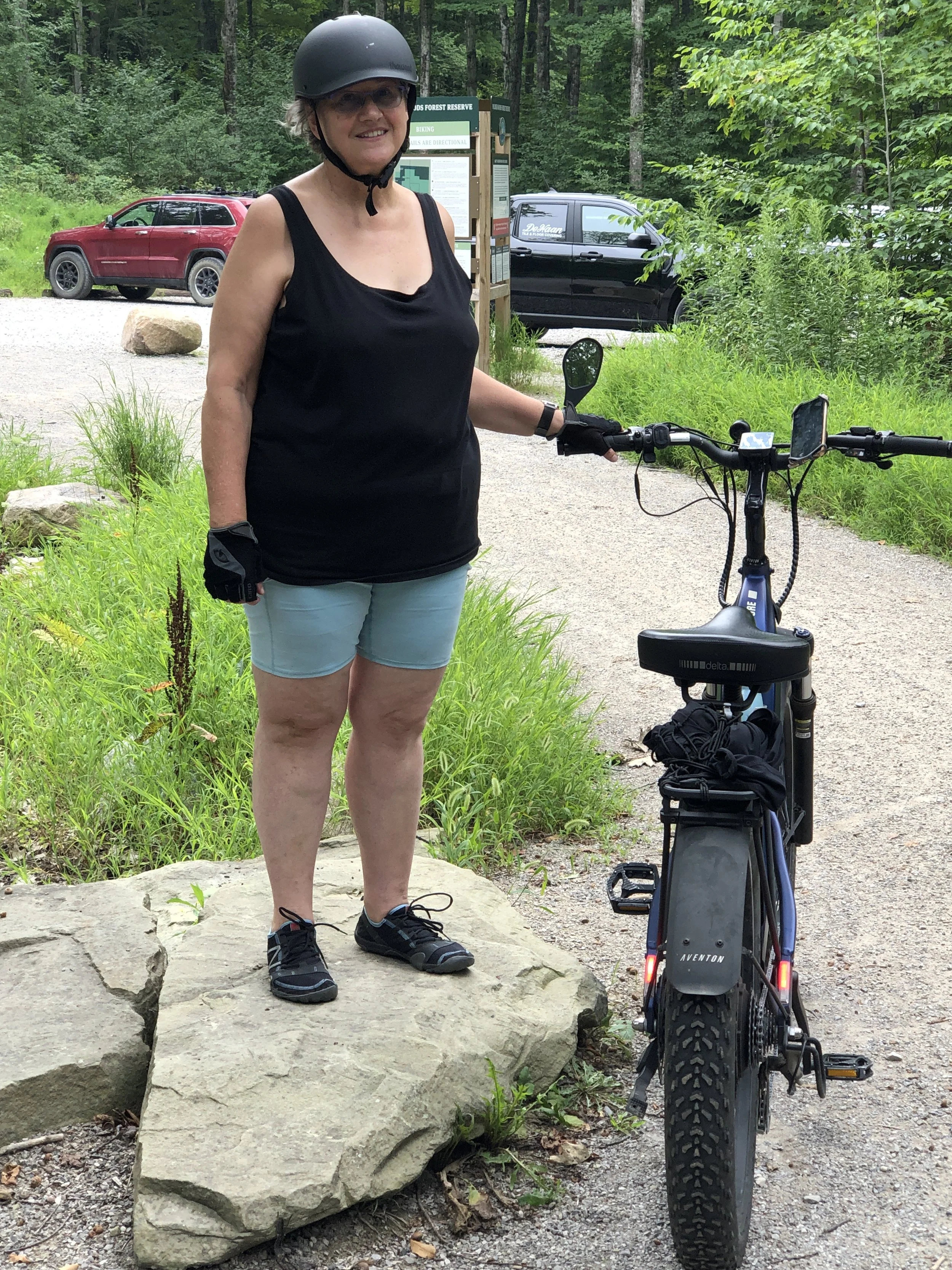 A woman wearing a black helmet, black tank top, and light blue shorts standing next to an electric bike on a gravel path in a green, wooded park.