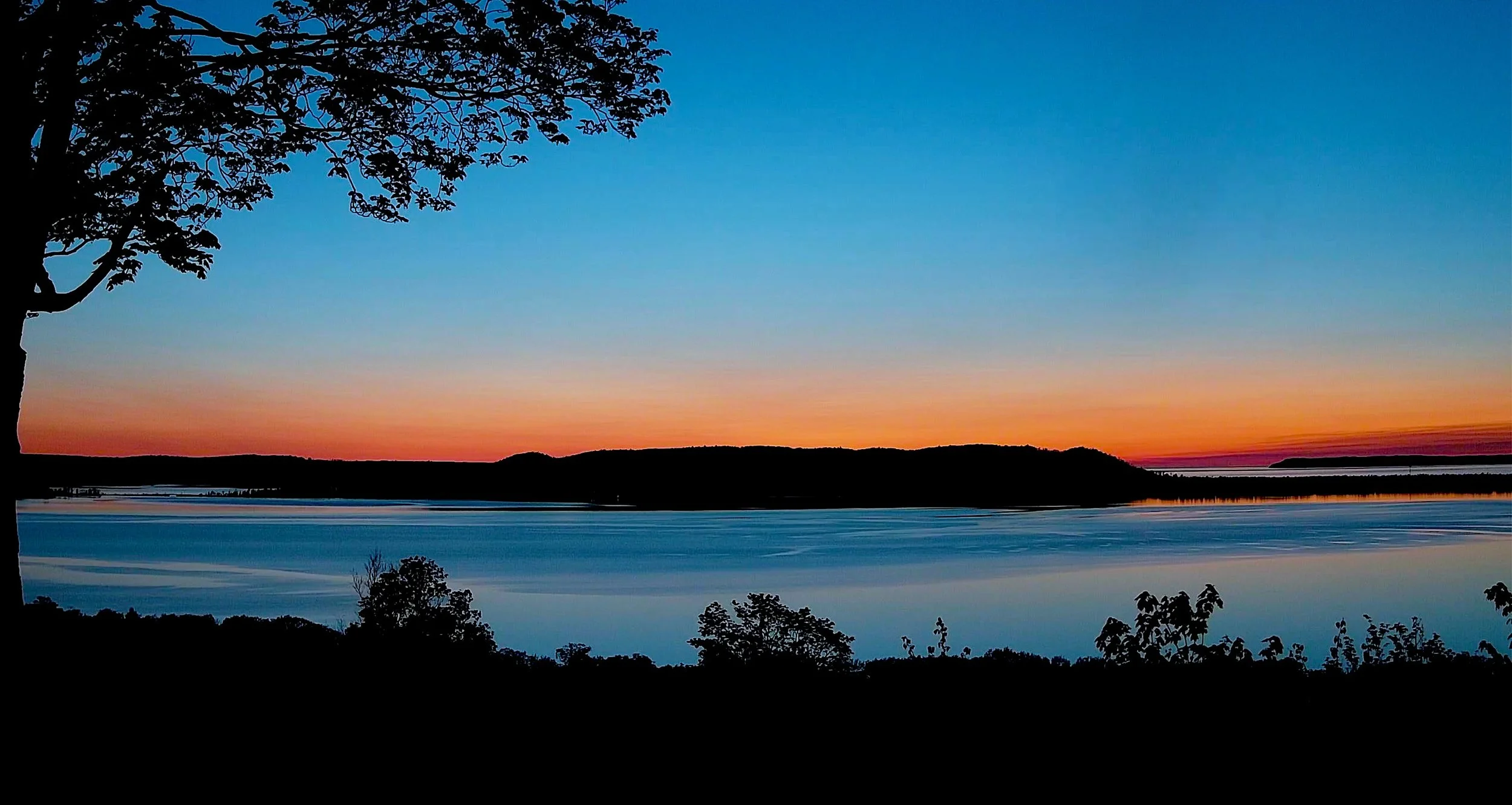 A scenic sunset over a large body of water with the sky changing from orange to blue and silhouetted trees in the foreground and along the horizon.