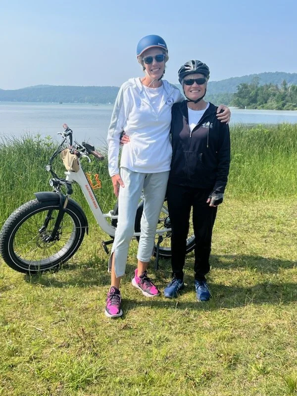 Two women standing outdoors on a grassy area near a lake, wearing helmets and sunglasses, with a black and white bicycle in the background. One woman is dressed in white with pink sneakers, and the other in black with blue shoes, smiling and posing t