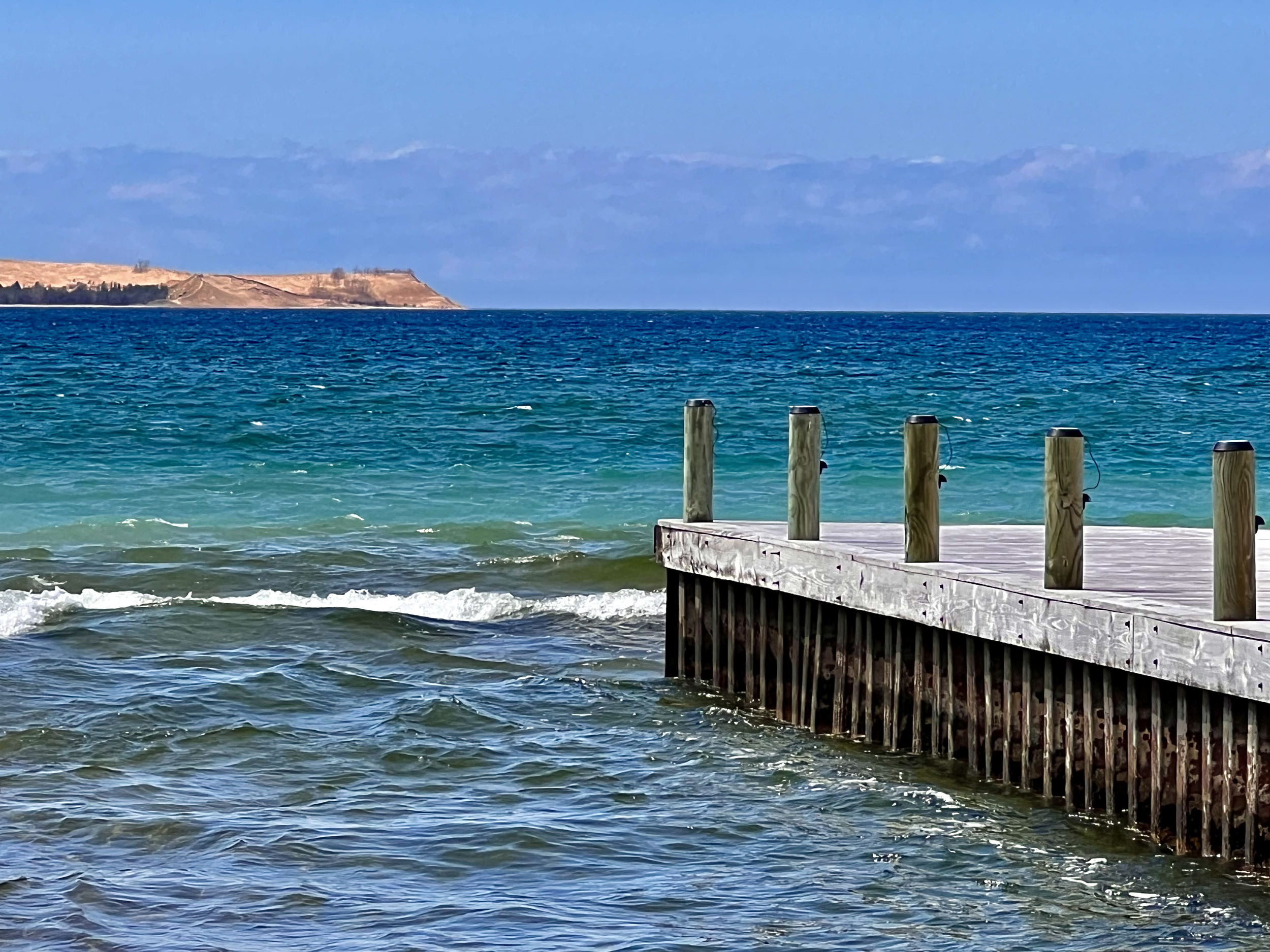 A wooden pier extends into a calm ocean, with small waves lapping against it. The water is a mix of blue and green hues. In the background, there is a landmass with cliffs and a partly cloudy sky.