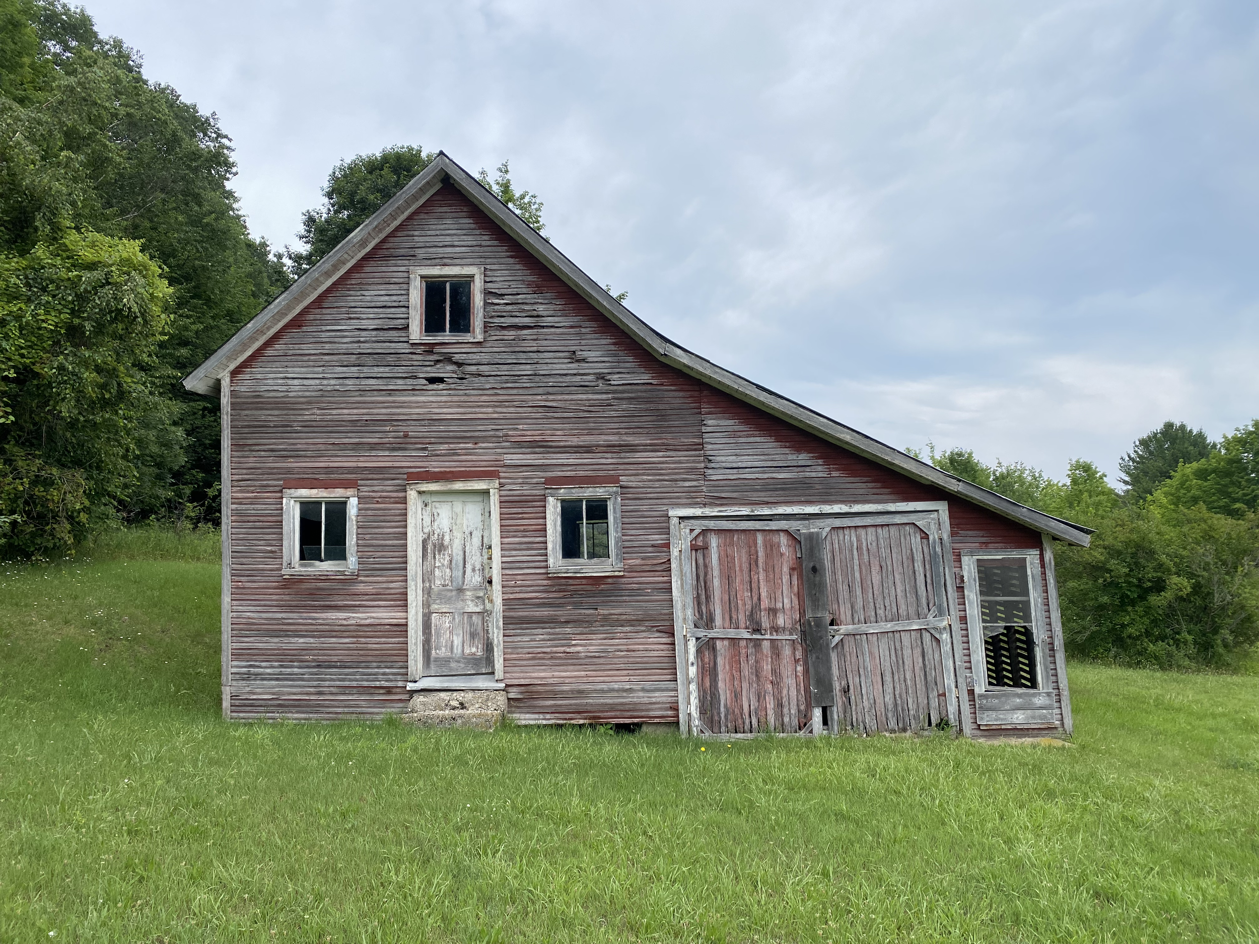 An old wooden barn with peeling red paint, worn-out windows, and a slightly tilted roof, surrounded by green grass and trees under a cloudy sky.