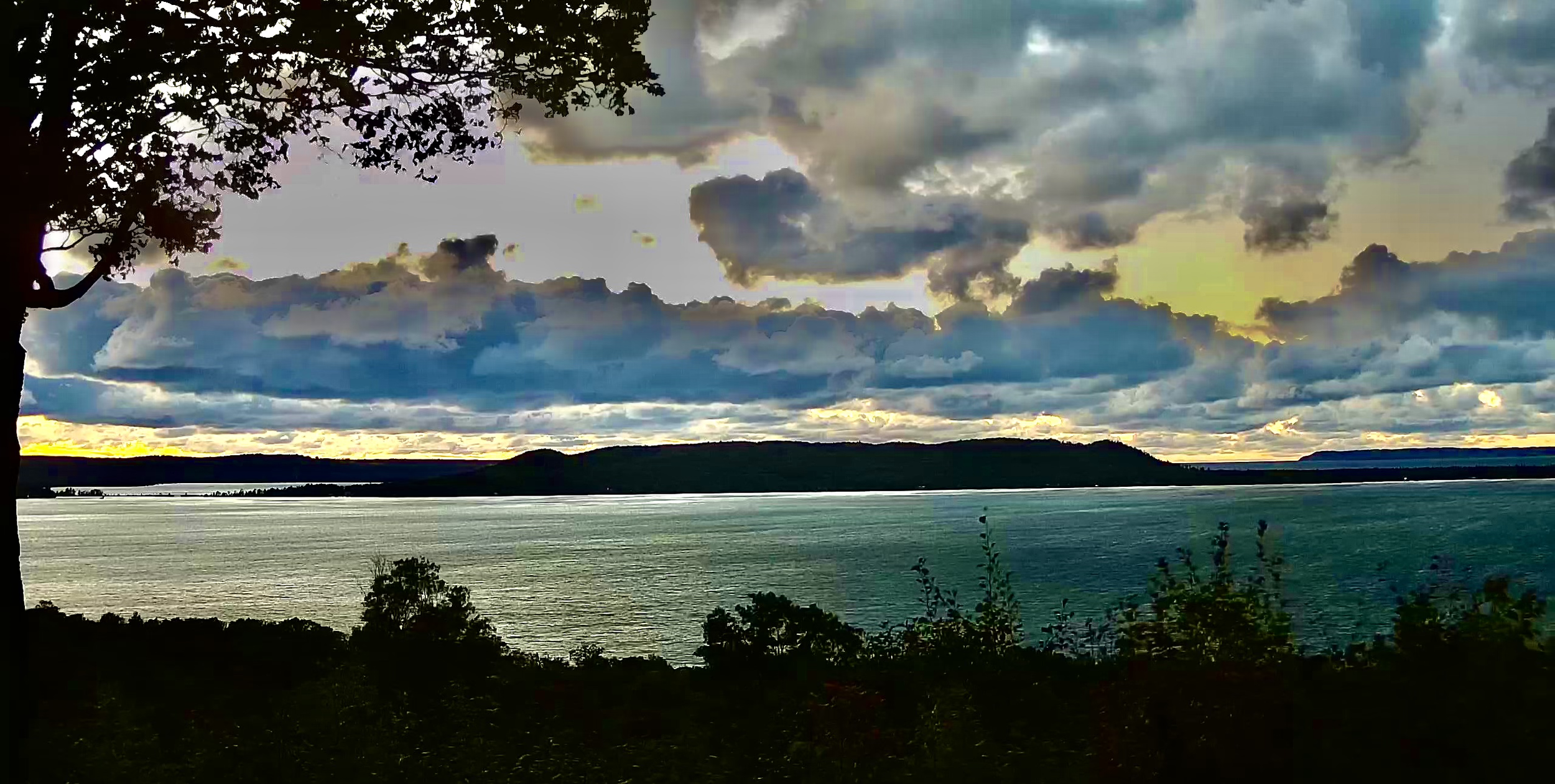 A view of a body of water with a tree branch and leaves in the foreground, dark hills in the distance, and a cloudy sky with hints of sunlight breaking through.