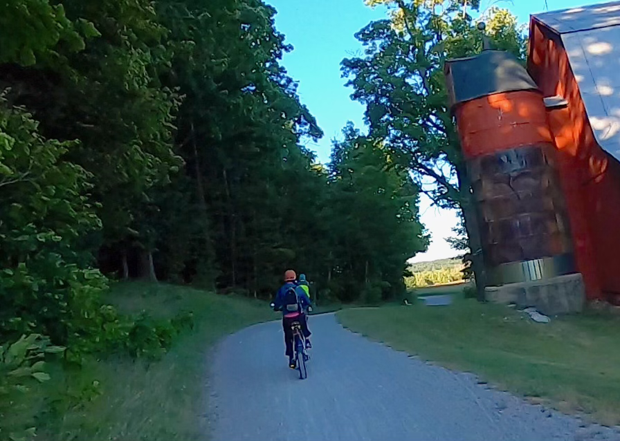 Two individuals riding bicycles on a gravel path through a wooded area near a large red barn. The children are wearing helmets and backpacks.