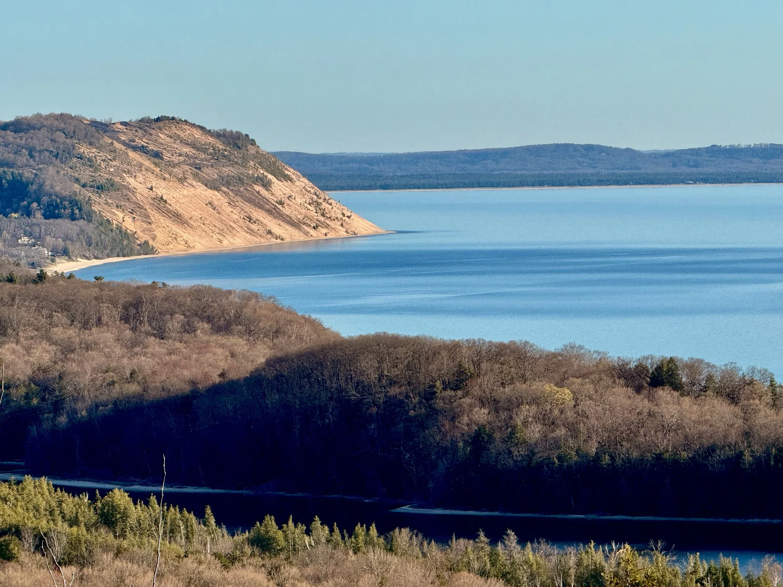 A lake with gentle ripples, surrounded by forests and rolling hills, under a clear blue sky.