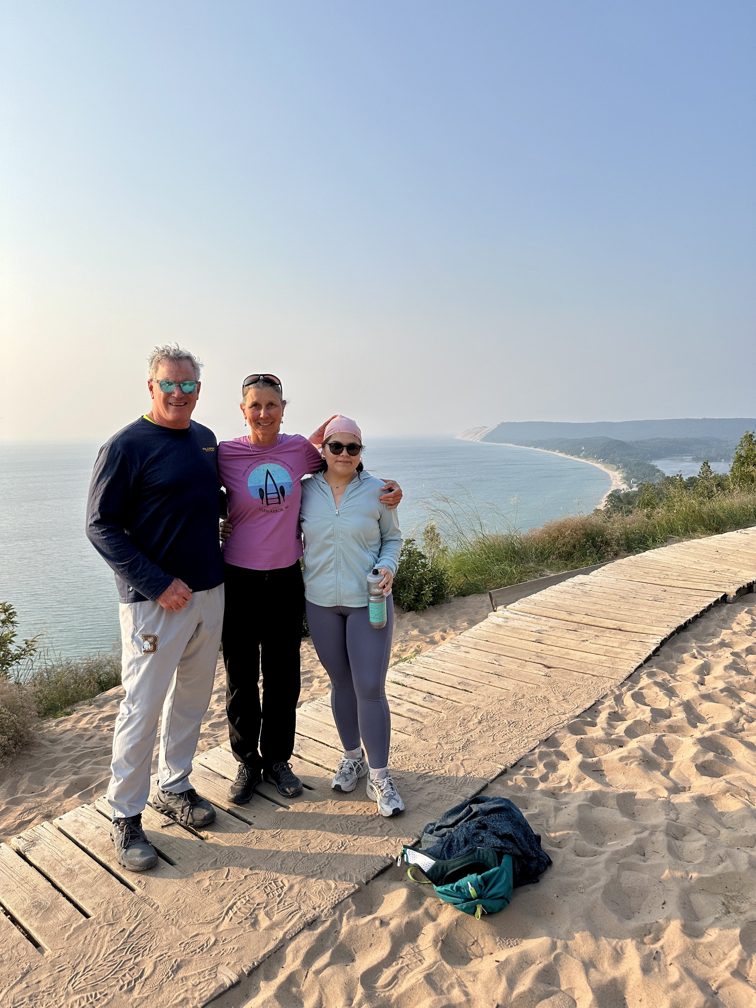 Three people standing on a sandy beach trail overlooking the ocean, with a distant shoreline in the background. They are dressed in casual and athletic clothing, smiling, with backpacks and water bottles nearby.