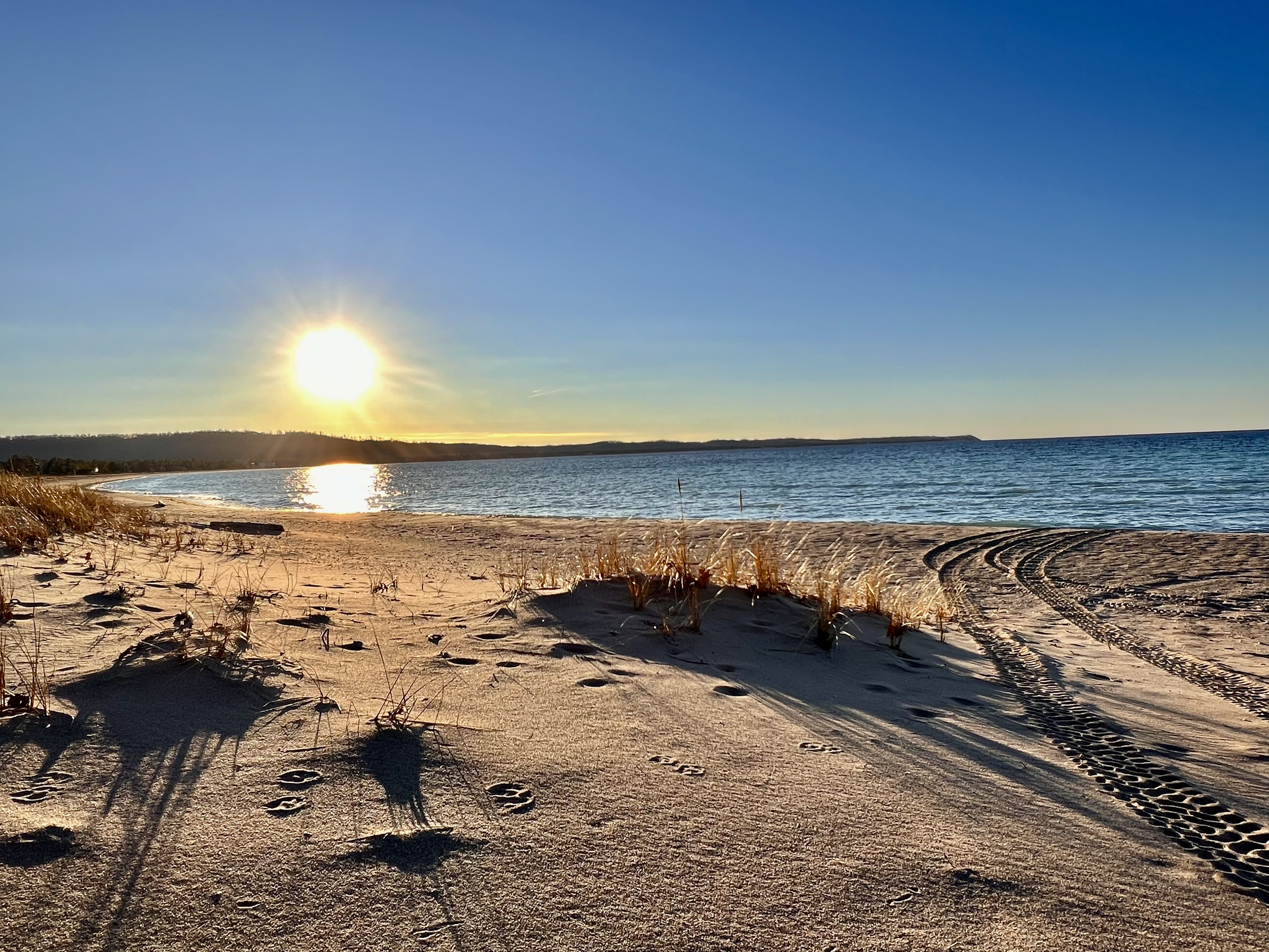 Sunset over a sandy beach with tire tracks, footprints, and sparse grass, facing a calm ocean.