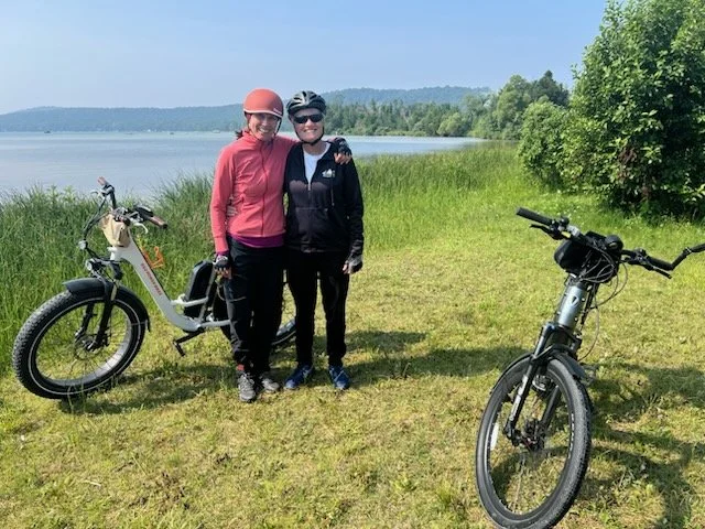 Two women standing on a grassy lakeside with two e-bikes, one woman wearing a pink jacket and helmet, the other in black with a helmet, with trees and water in the background.