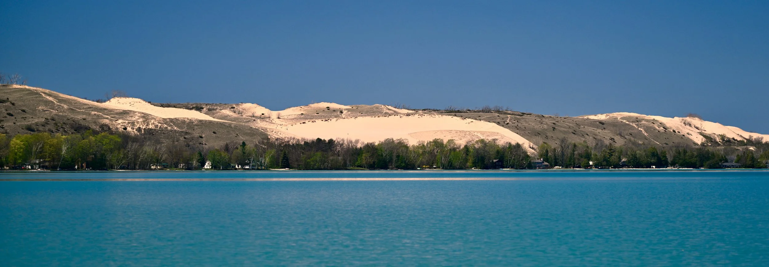 A scenic view of a lake with turquoise water, green trees along the shoreline, and a large hill with sandy patches and sparse vegetation under a clear blue sky.