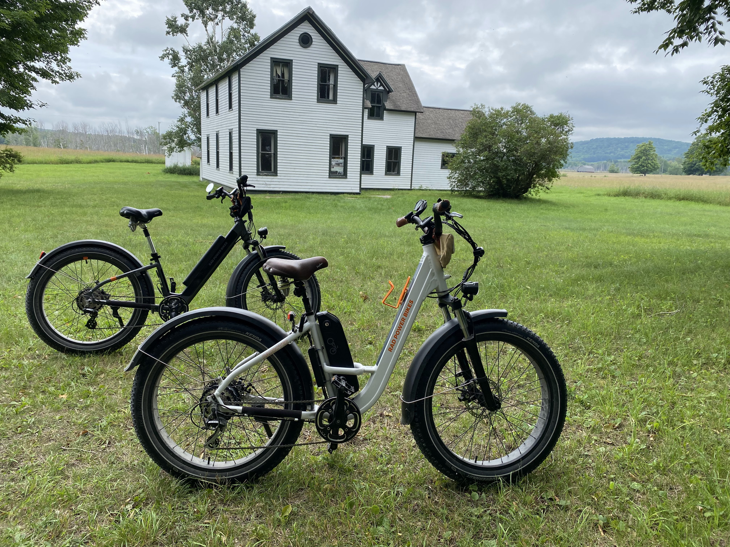 Two electric bikes on a grassy field with a white, two-story house and trees in the background.