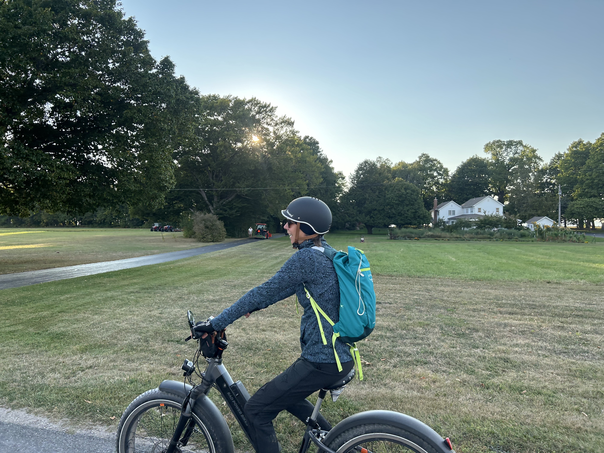 A woman wearing a helmet and sunglasses riding a bicycle outdoors on a sunny day, with a residential area and large trees in the background.