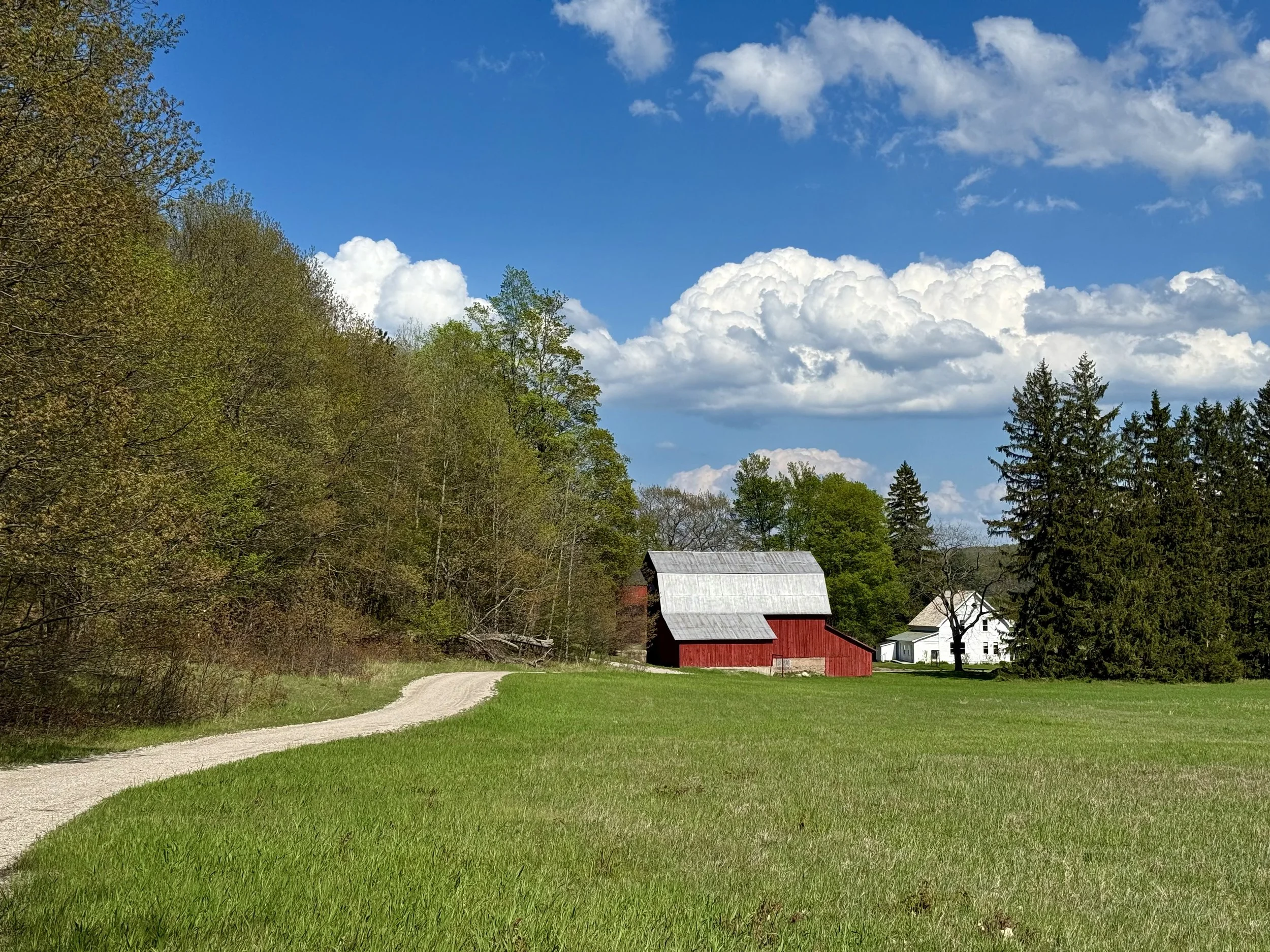A rural landscape with a grassy field, a winding dirt road, a red barn, and a white house surrounded by trees under a partly cloudy blue sky.