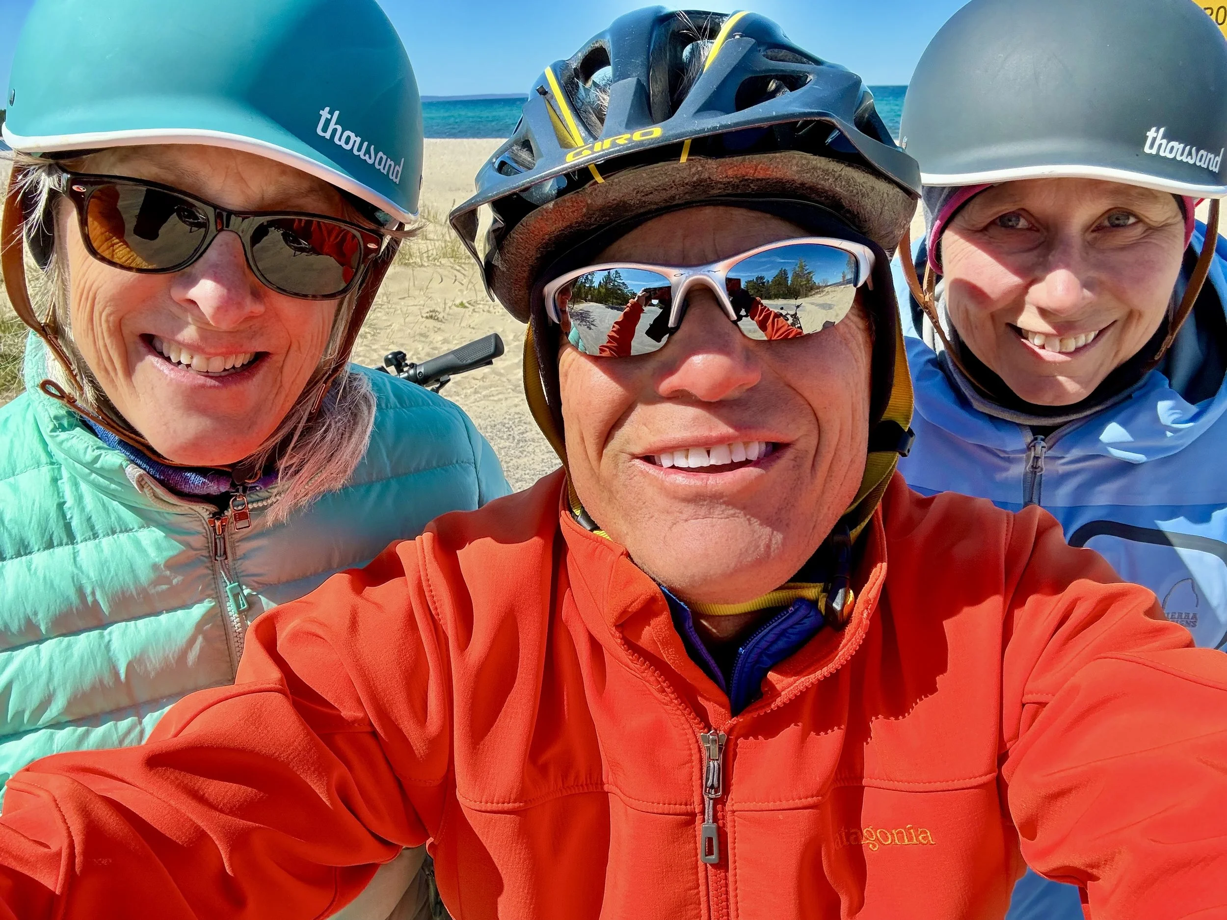 Three people wearing helmets and sunglasses taking a selfie at the beach with sand and ocean in the background.