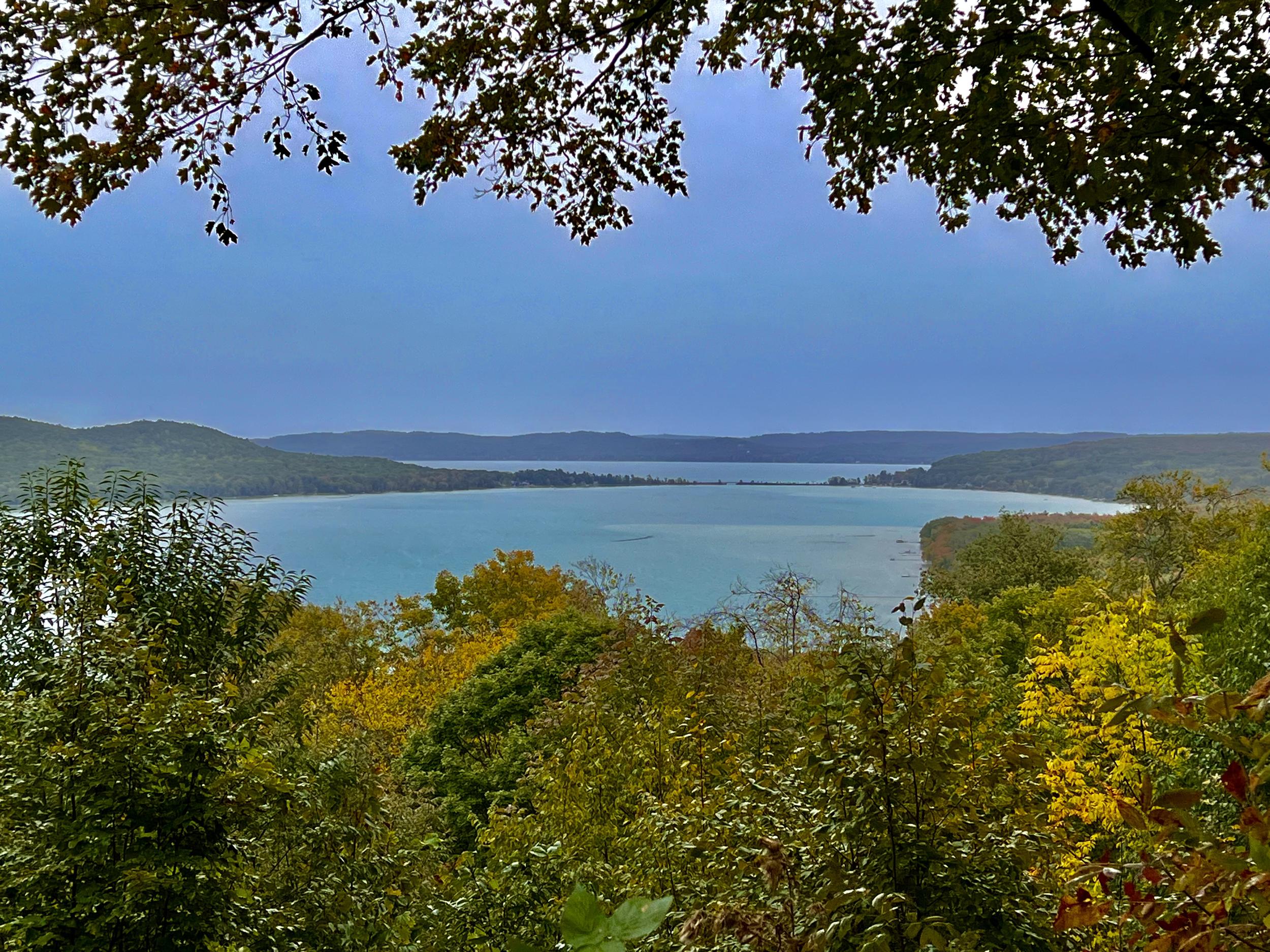 A scenic view of a lake surrounded by trees with green and autumn foliage, with hills in the background and a partly cloudy sky overhead.
