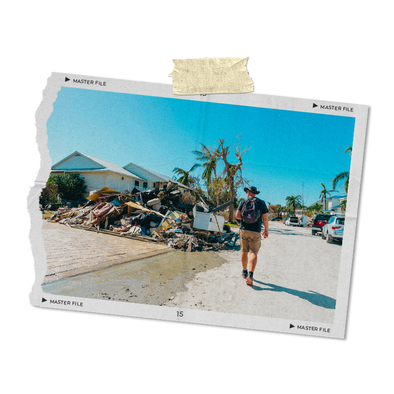 A person with a backpack and hat walking away on a street after a storm, with debris and damaged trees, surrounded by houses and palm trees under a clear blue sky.