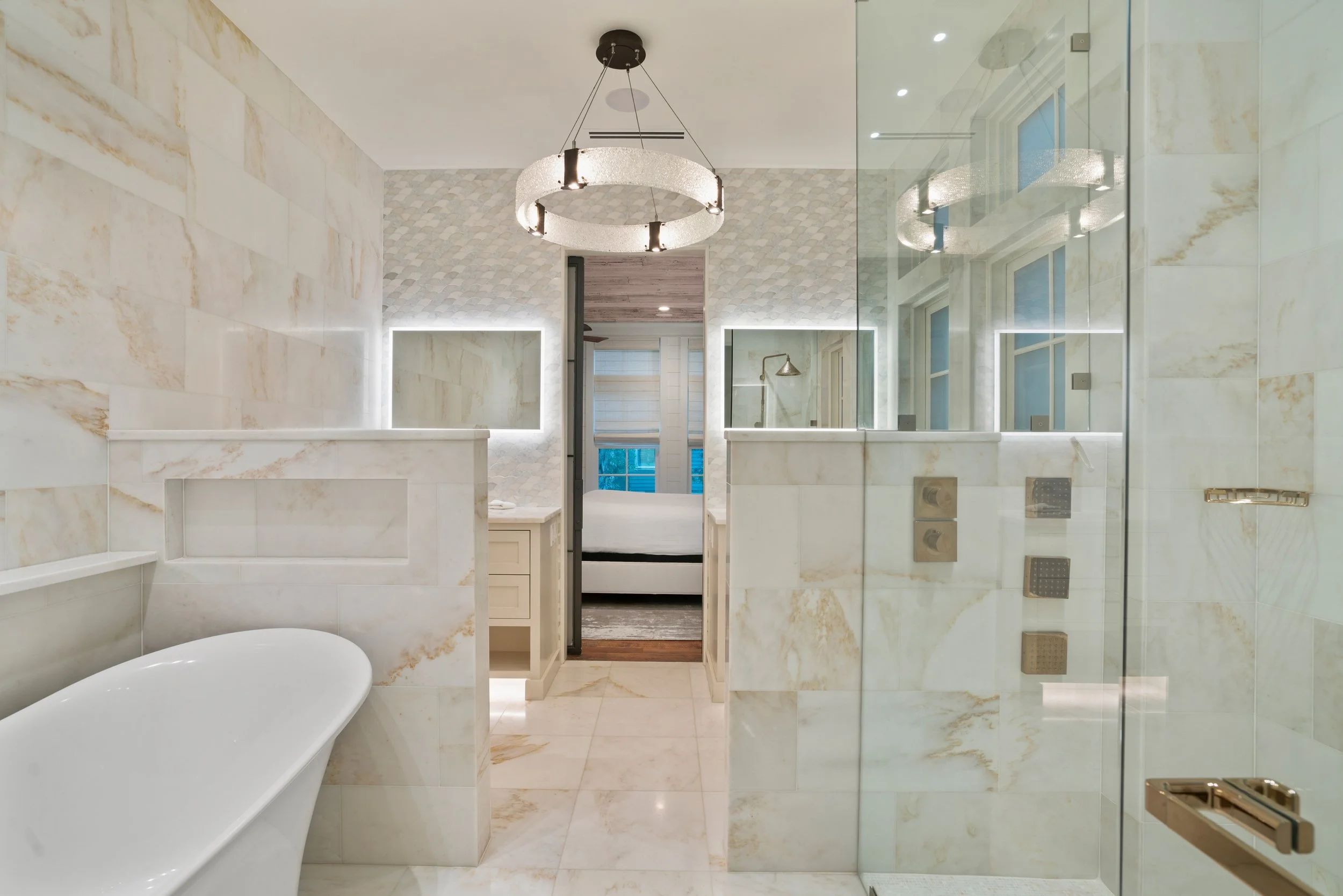 Modern bathroom with marble tiles, a bathtub, glass shower enclosure, double sinks with illuminated mirrors, and a view of a bedroom with white bedding through a doorway.
