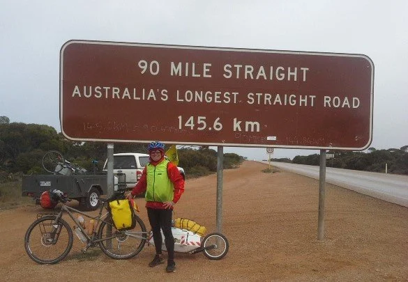 Cyclist in helmet and bright clothing standing beside a loaded bicycle and Quest bike trailer near a large road sign