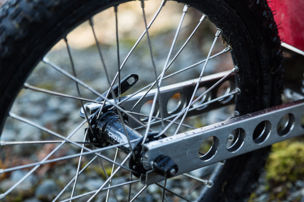 Close-up of a Quest bike trailer wheel with spokes and a rear hub, part of the bike frame, and a rocky ground background.