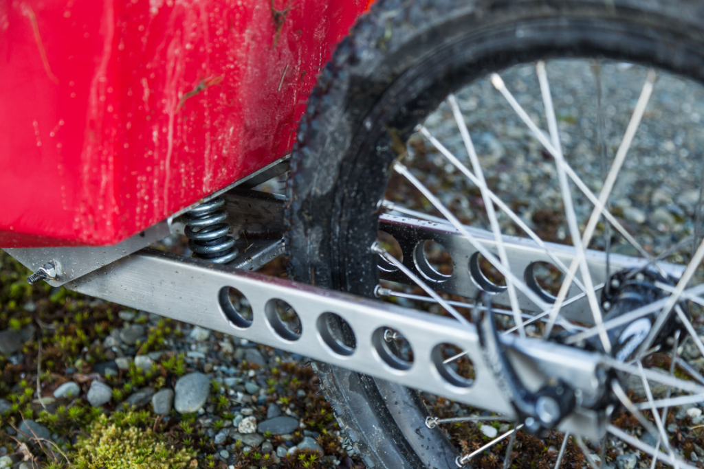 Close-up of a Quest bike trailer wheel with a black tire and spokes, attached to a frame with a visible spring, on a gravel and moss-covered ground.
