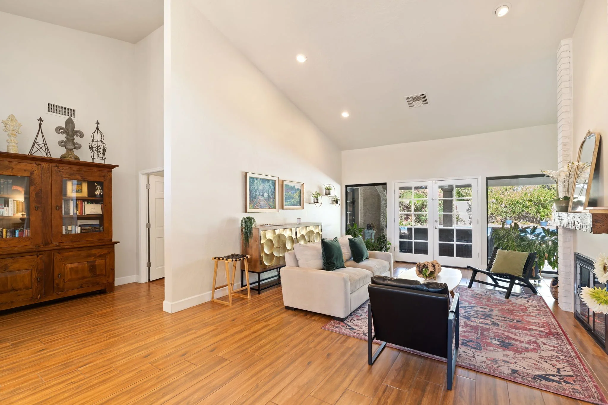 Bright living room with white walls, hardwood floors, a white sofa with green pillows, a black armchair, a patterned area rug, a wooden bookshelf, a gold sideboard, a fireplace, and large glass doors leading to an outdoor patio.