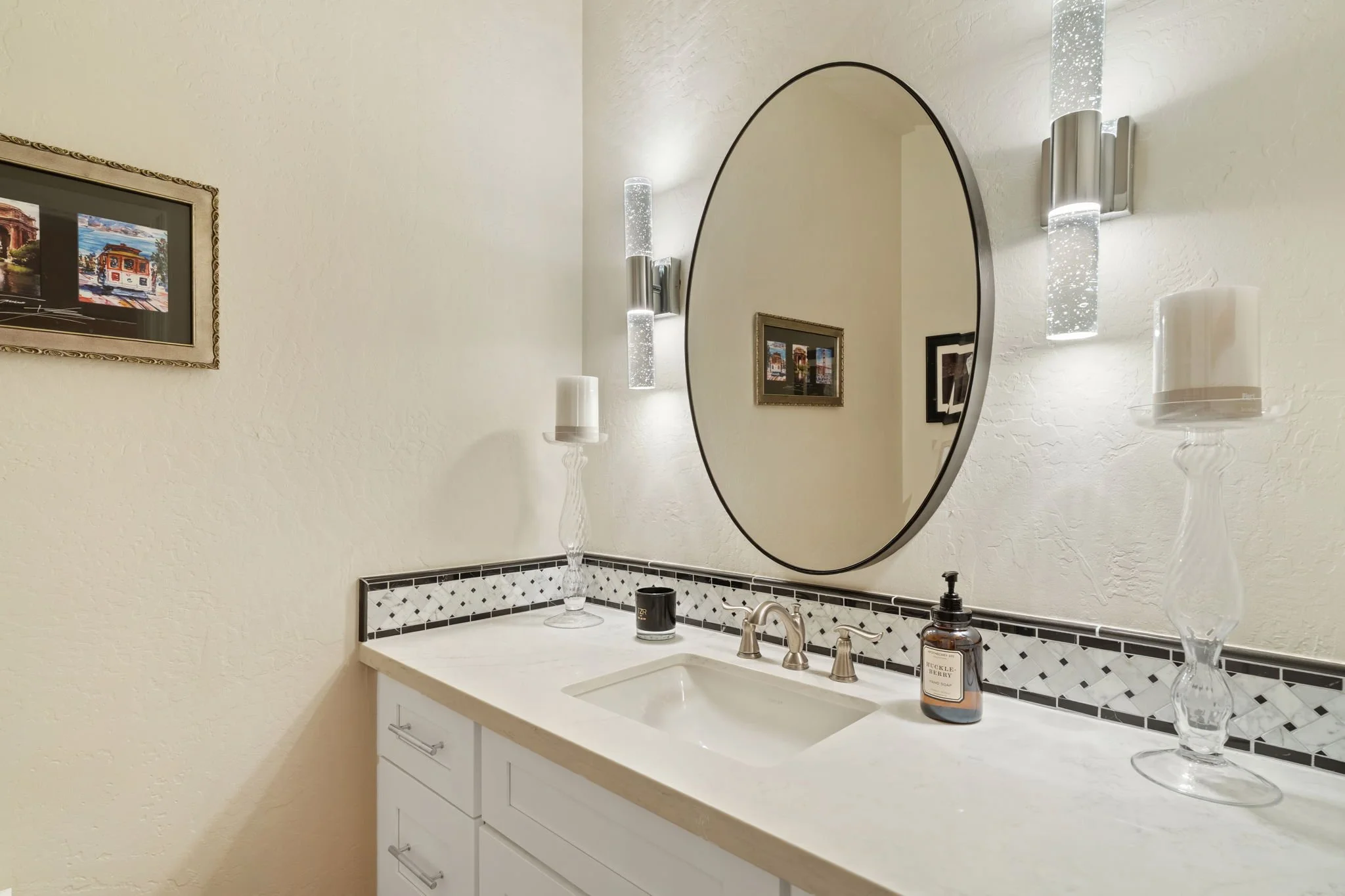 Bathroom sink vanity with a large oval mirror, two decorative glass candle holders, a soap dispenser, and framed pictures on the wall.