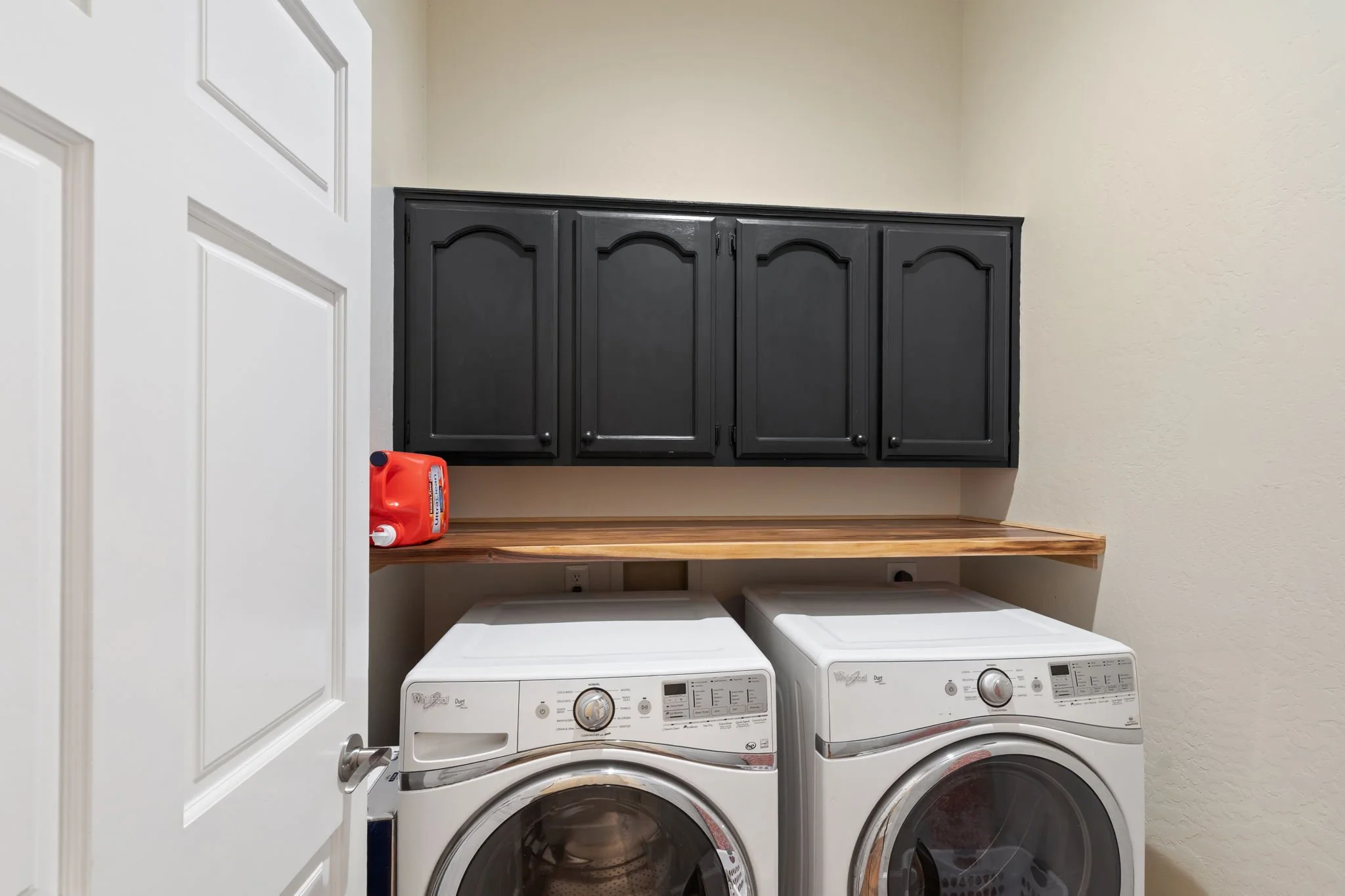Laundry room with white washer and dryer, black cabinets, wooden shelf, and a bottle of laundry detergent.