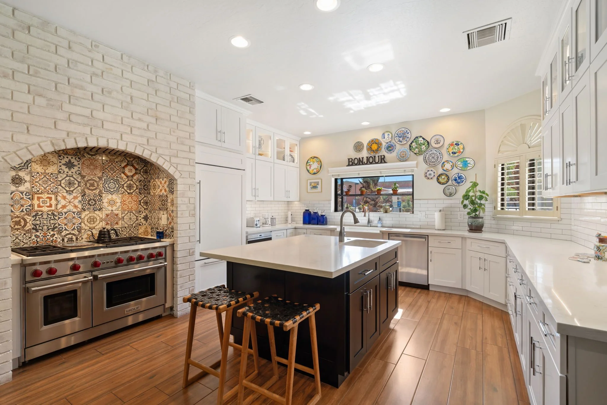 Modern kitchen with white cabinets, a black kitchen island, tiled backsplash, stainless steel appliances, a window with a 'Bonjour' sign and decorated plates on the wall, and wood flooring.