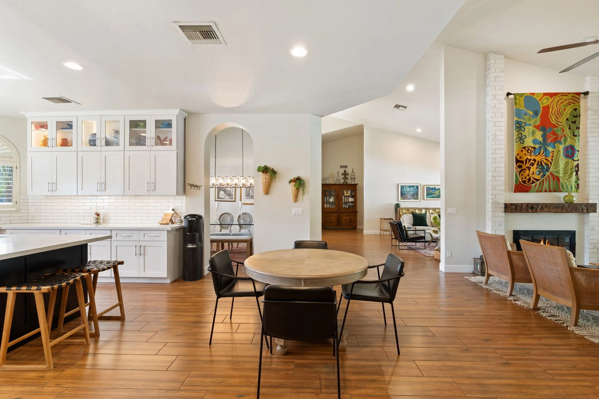 Open-concept living space with white cabinetry, a round dining table, black chairs, wooden flooring, a fireplace with colorful art, and a cozy seating area with additional chairs and artwork.