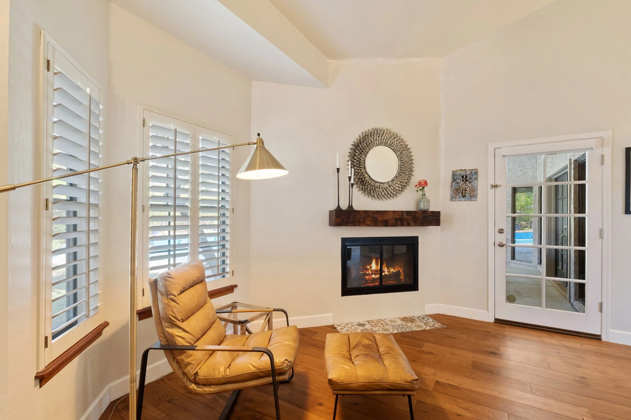 Living room with a fireplace, wooden mantel, a round mirror, a beige lounge chair with matching ottoman, wooden floors, and a glass door leading outside.