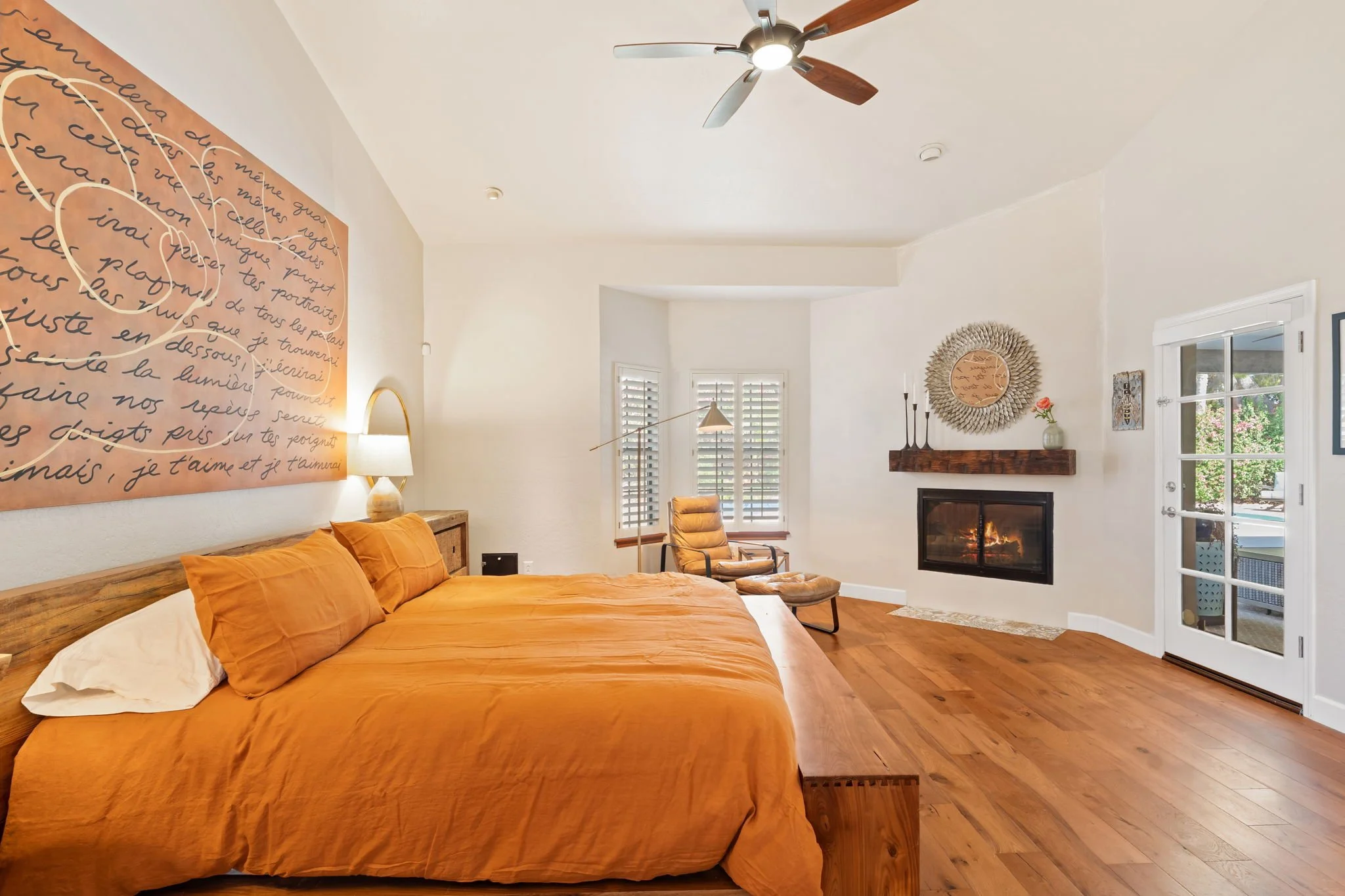 Primary bedroom with a wooden bed frame, orange bedding, a bedside lamp, a sitting chair near windows with white shutters, a fireplace with a decorative mirror and a door leading outside. The room has a wood ceiling fan and hardwood floors.