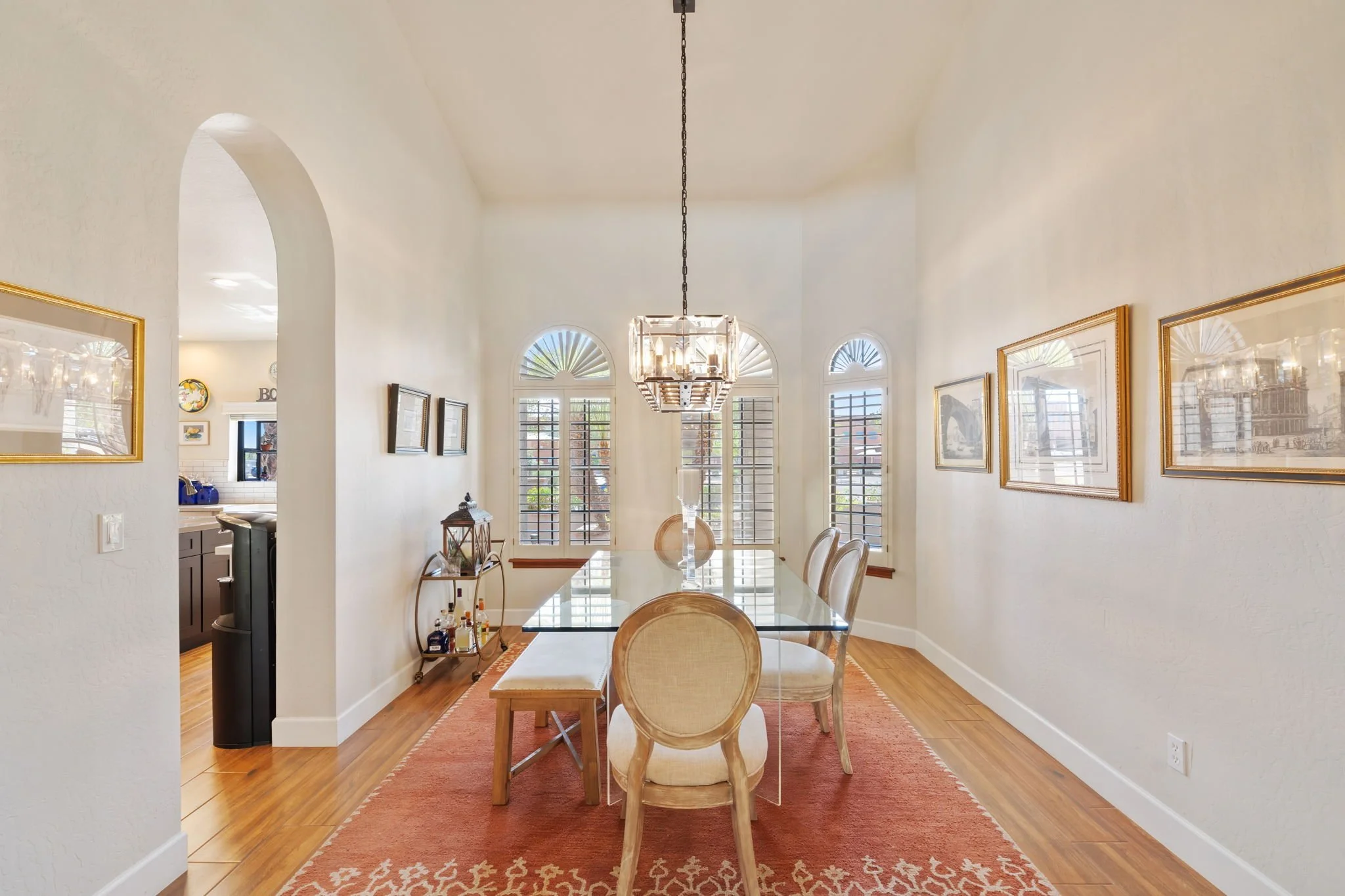A dining room with a glass-top table, six upholstered chairs, and a chandelier hanging above. The room has large arched windows with shutters, framed artwork on the walls, and a red area rug on wooden flooring.