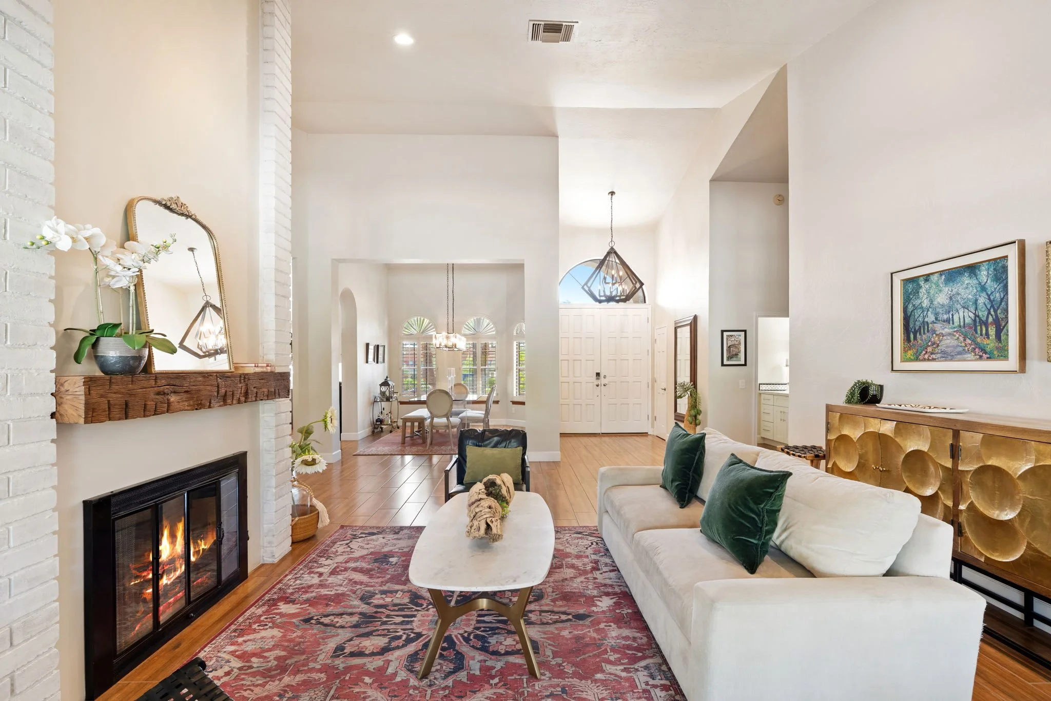 Living room with white walls and high ceilings, a fireplace with a wooden mantel, a cream sofa with green pillows, a marble coffee table, a patterned area rug, and artwork on the walls. A dining area with a chandelier and a round table is visible in