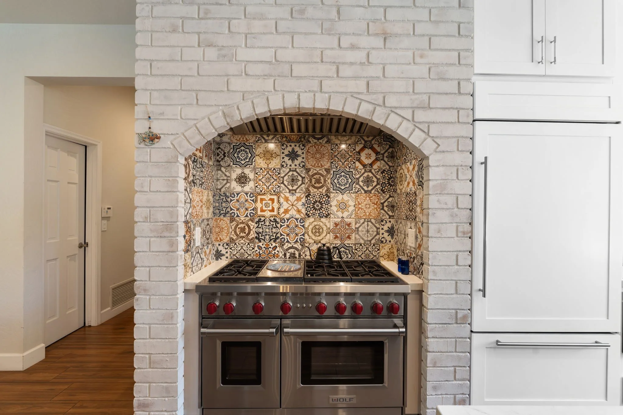 Kitchen with a white brick archway framing a stainless steel Wolf subzero stove and oven with red knobs, backed by a colorful patterned tile backsplash, next to white cabinetry.