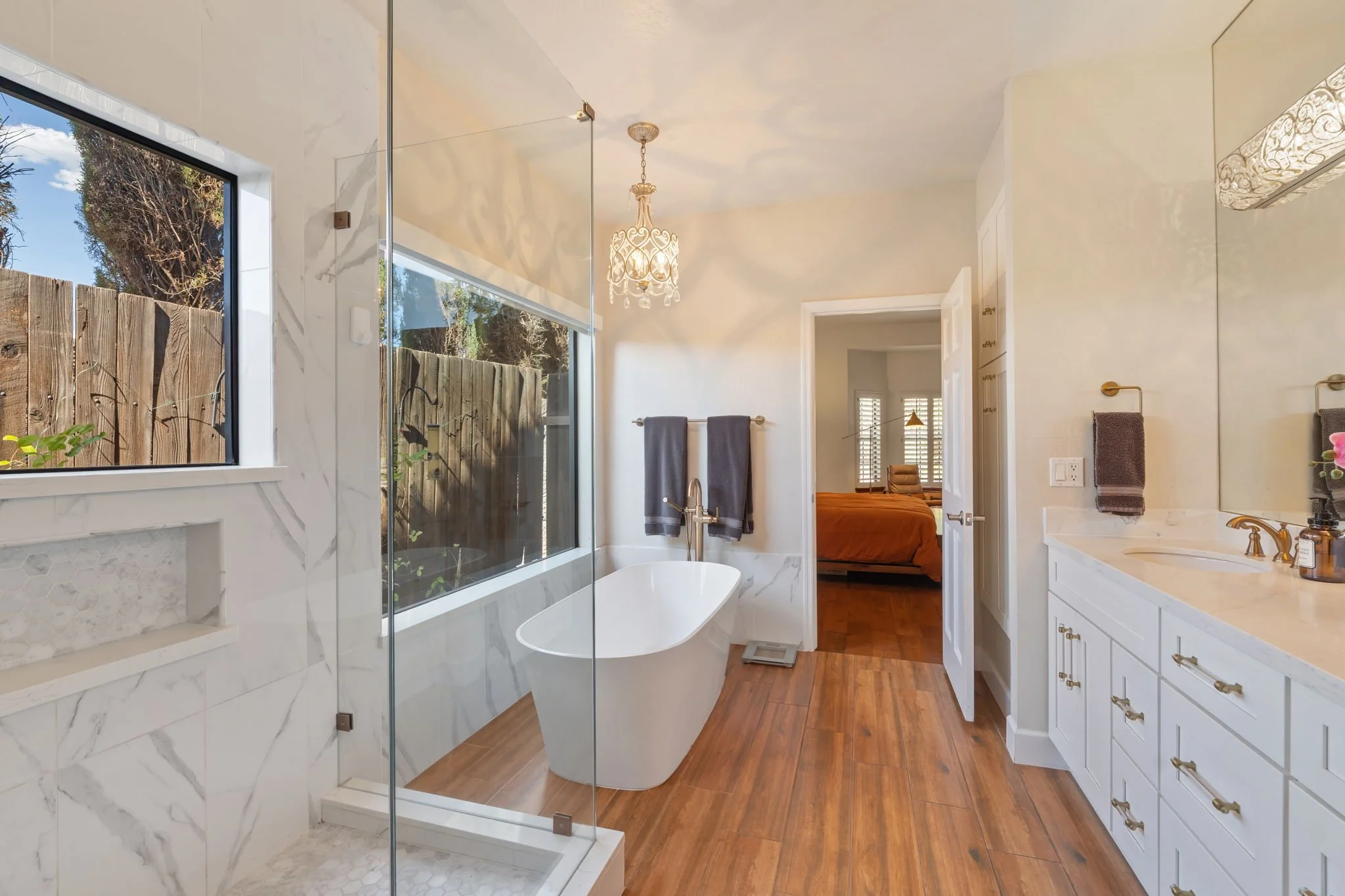 Modern bathroom with free-standing bathtub, glass shower enclosure, large window, wooden flooring, white vanity with gold fixtures, and a view into the primary bedroom.
