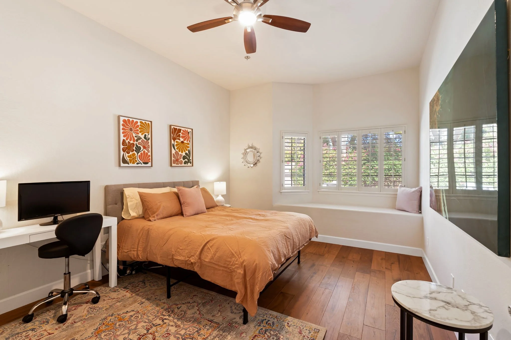 A bedroom with a bed, nightstand lamps, framed art, a small desk with a computer, and a window with white plantation shutters.