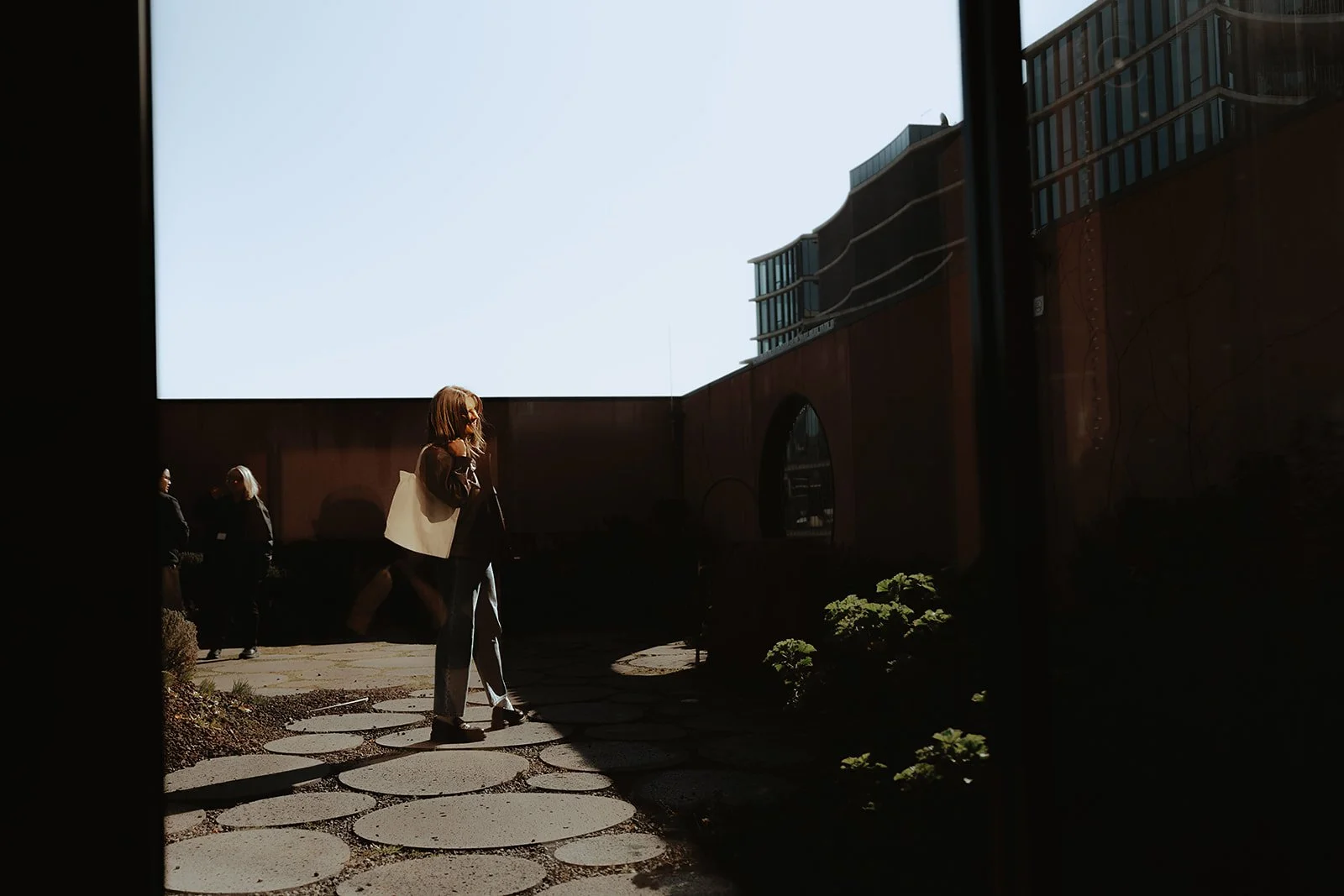 Person standing on a terrace rooftop outside, with two other people slightly blurred in the background. Bright sunlight creates shadows, and the sky is clear and blue.