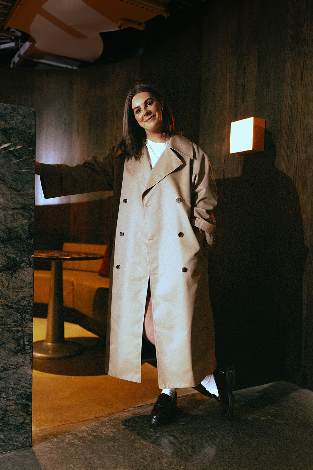A woman in a beige trench coat standing in an office and leaning against a wall, smiling softly.